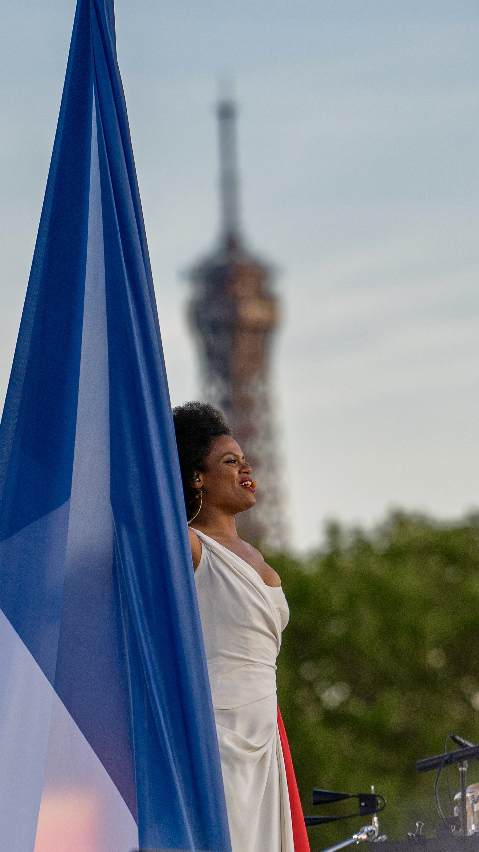 Une femme africaine en robe blanche, souriant, se tient à côté d'un drapeau bleu et blanc en extérieur, avec une tour en arrière-plan.