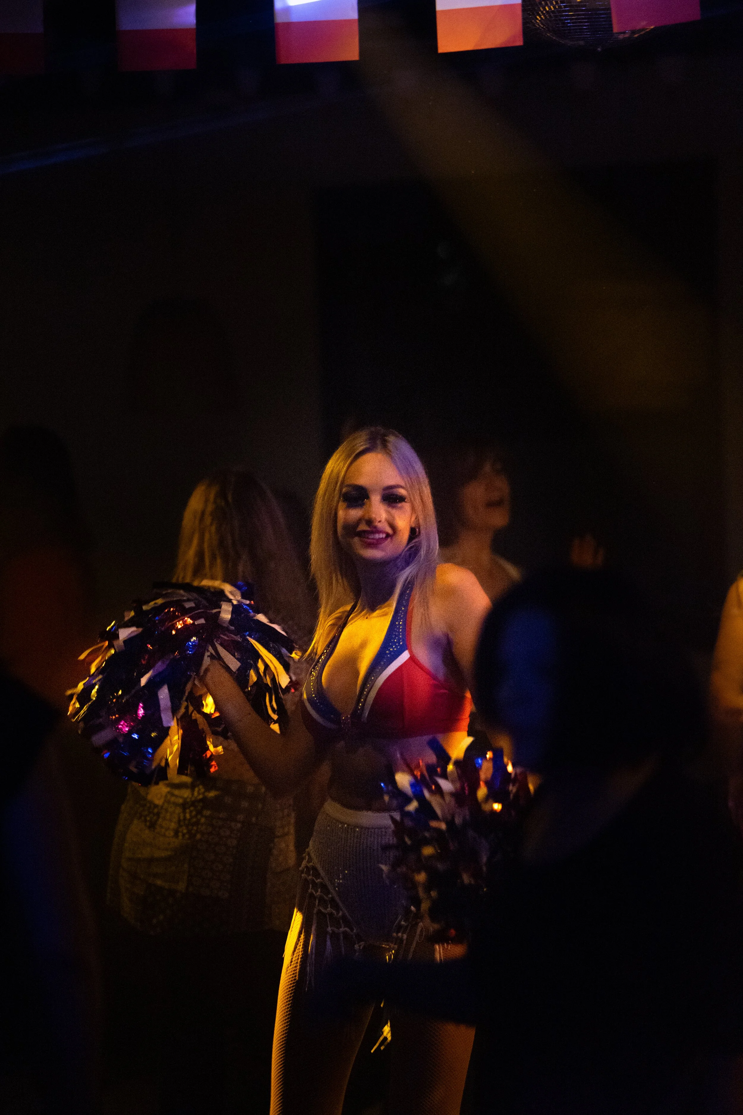 Une danseuse de cheerleading vêtue d'un costume coloré en rouge, blanc et bleu, tenant des pompons colorés, dans une ambiance sombre avec des lumières de scène. Elle sourit à l'appareil, entourée d'autres personnes.