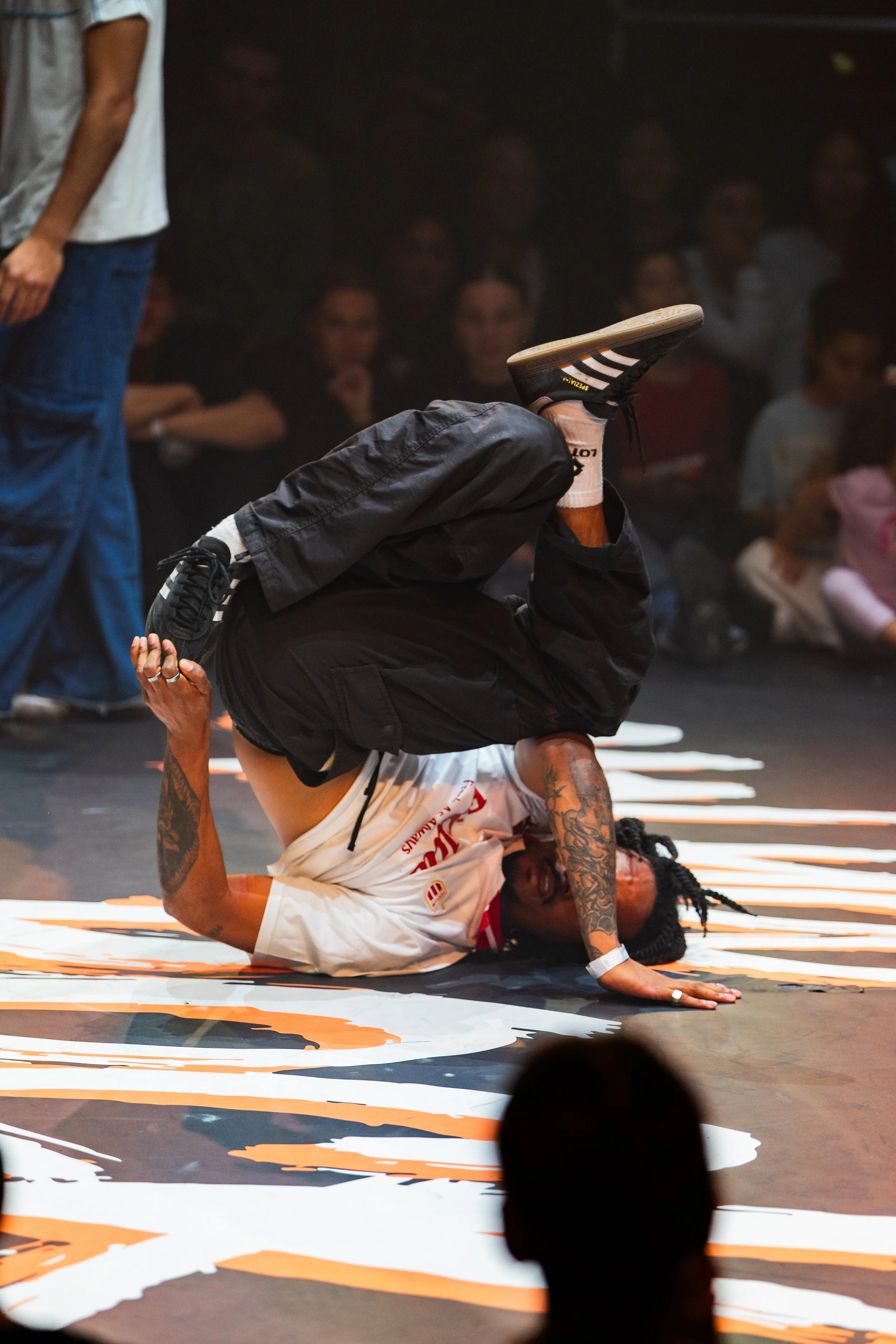 Un danseur de breakdance effectuant un mouvement acrobatique au sol lors d'une compétition, avec un public en arrière-plan.