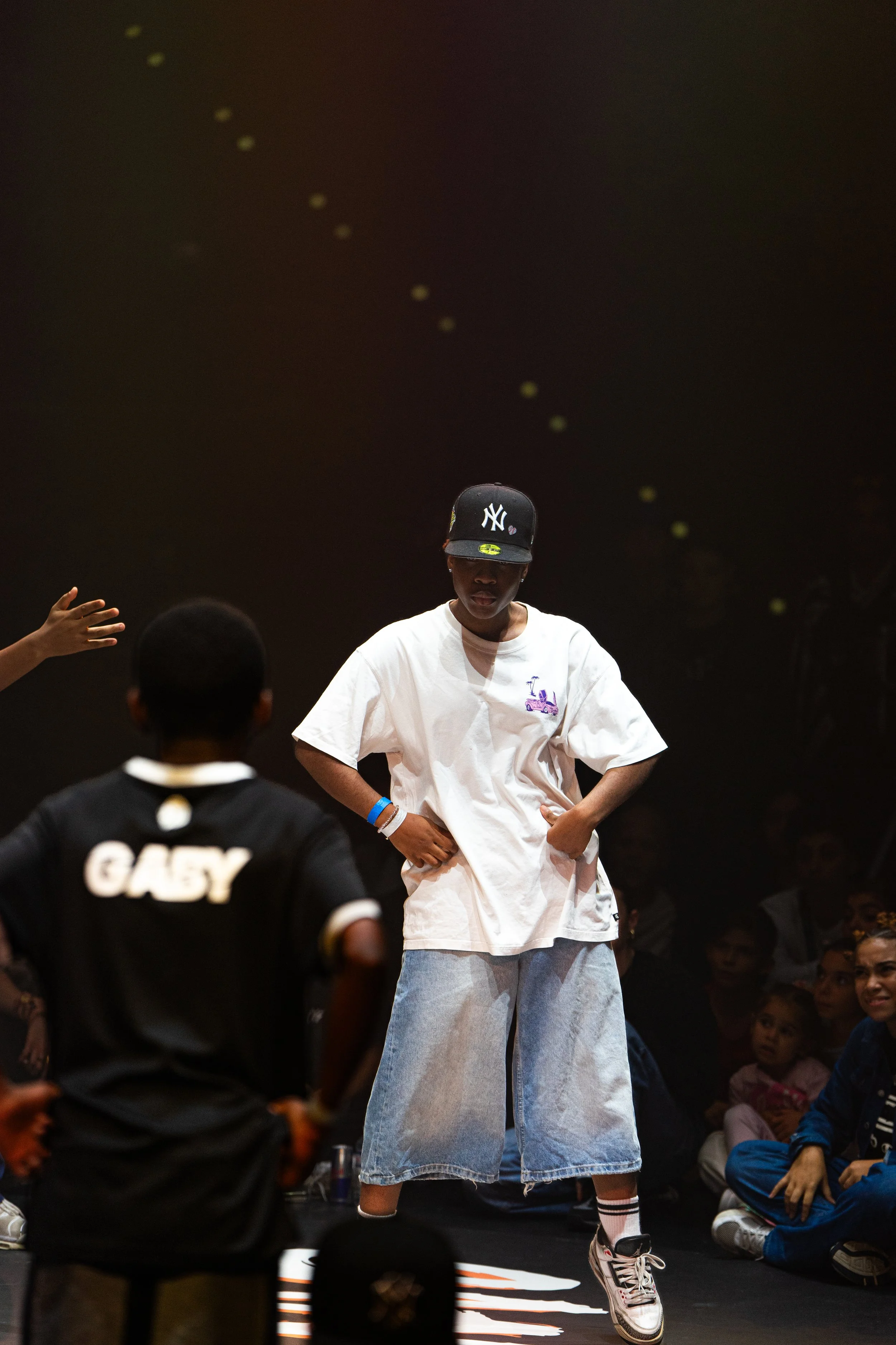 Une personne en vêtements décontractés, portant une casquette noire avec le logo des Yankees, se tient sur scène avec une main sur la hanche, entourée d'un public, dans ce qui semble être une scène de danse ou de performance.