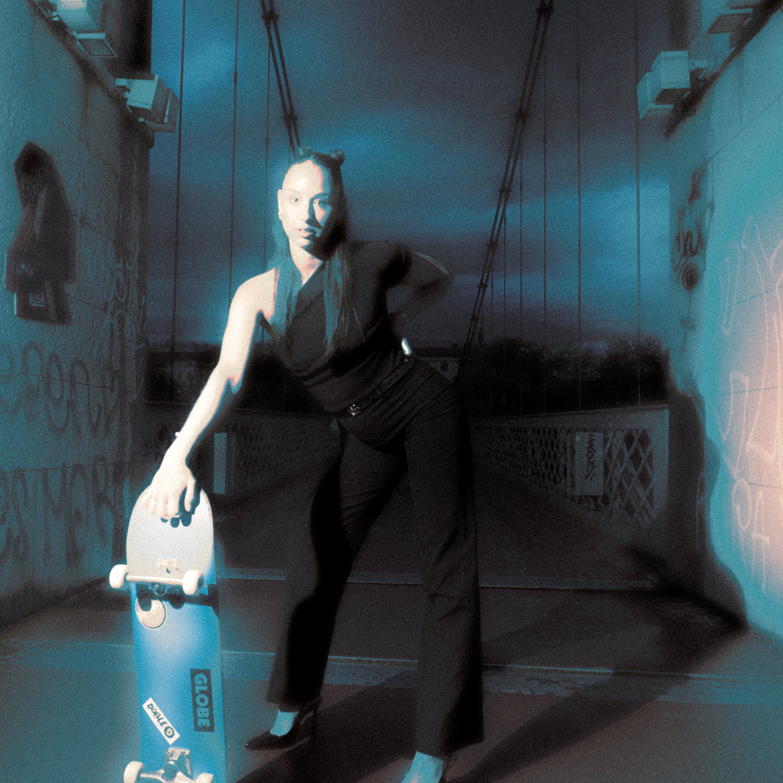 Une femme avec des cheveux longs coiffés en deux petits buns, portant une tenue noire, pose avec une planche à roulettes dans un endroit sombre avec des graffitis et des graffiti sur le mur, à la lumière bleue.