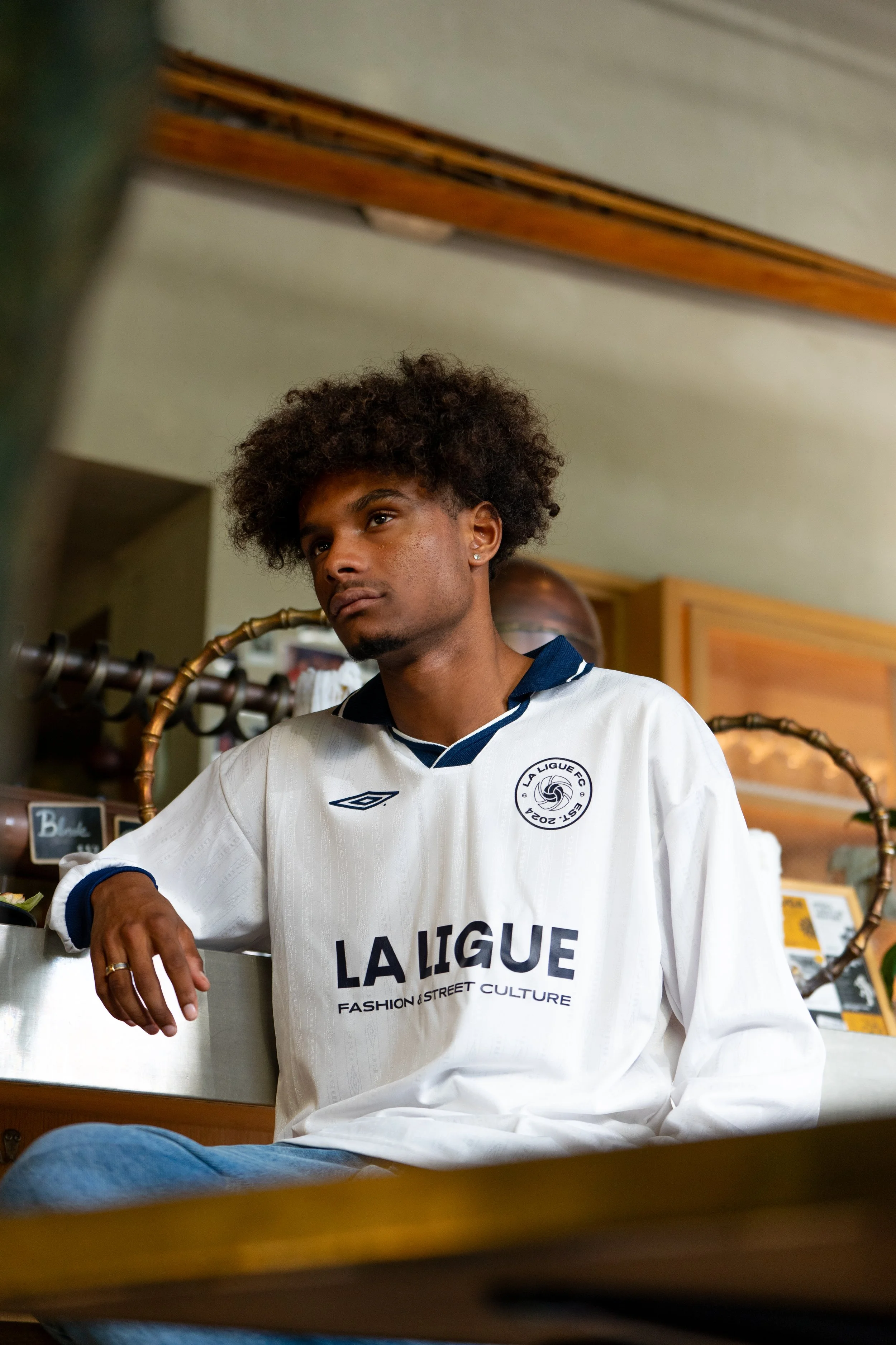 Jeune homme assis, portant un maillot blanc de La Ligue, avec un logo de football, dans un espace intérieur.
