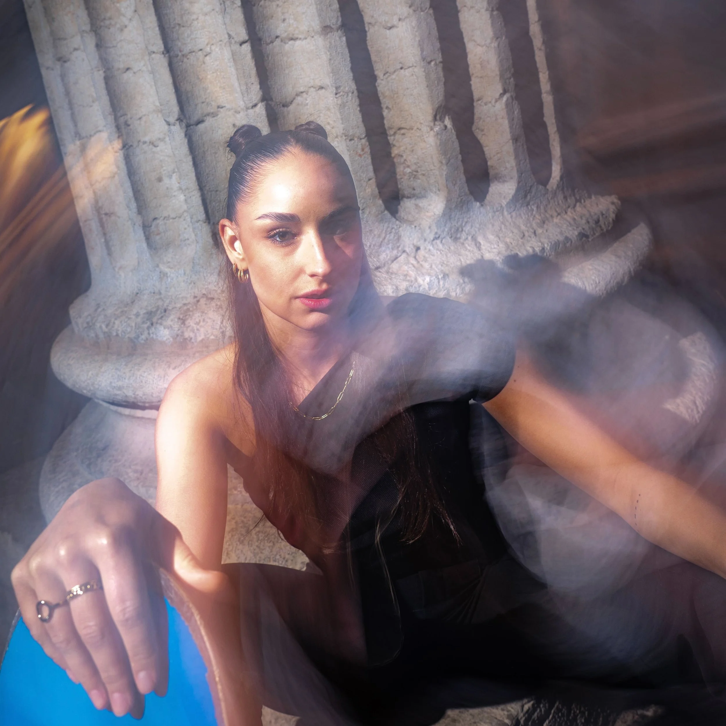 Une femme assise devant une colonne antique en pierre, portant une robe noire et des bijoux dorés, avec une lumière douce et un effet de flou artistique.