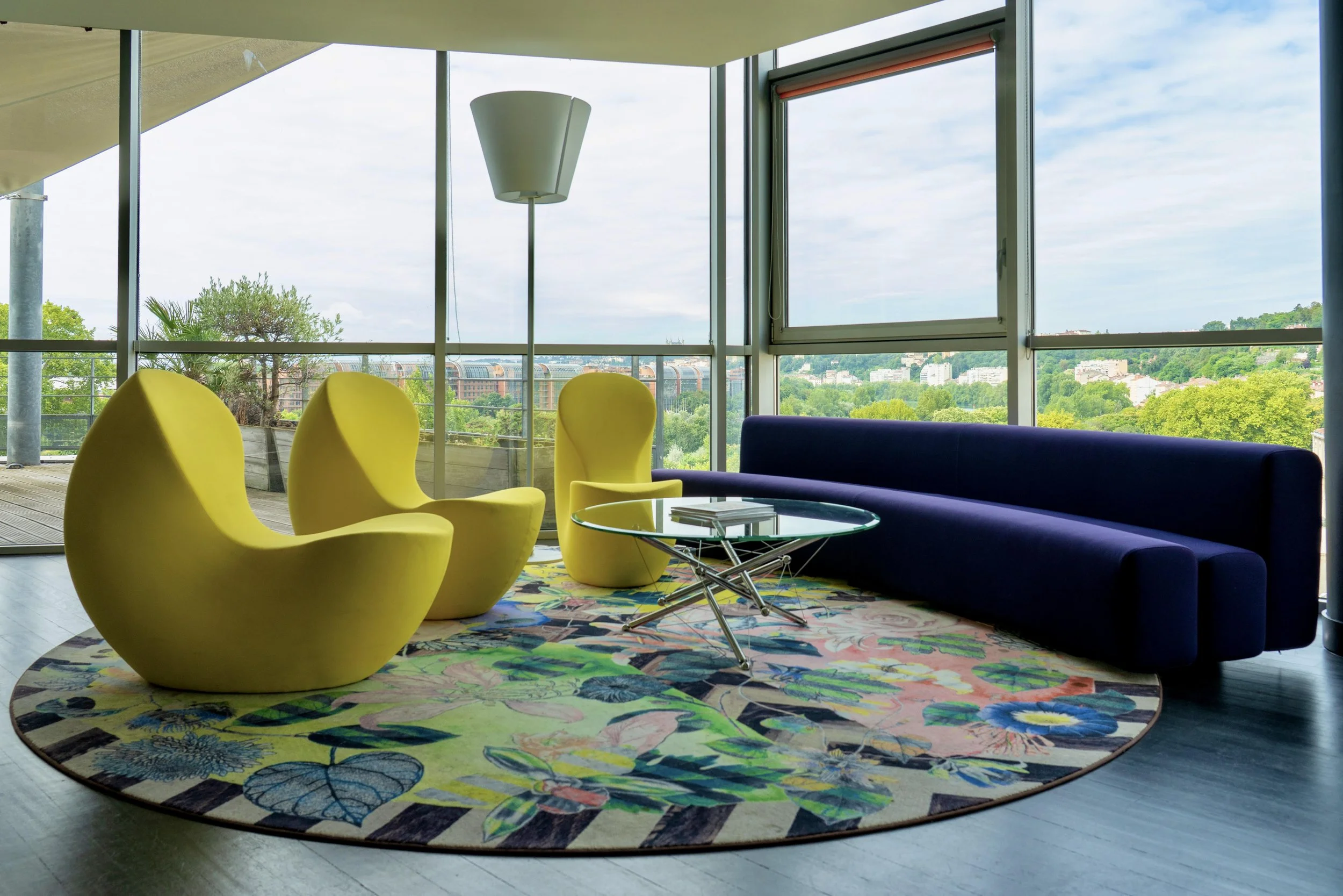 Salon avec trois chaises jaunes modernes, un canapé bleu foncé, une table en verre ronde, un tapis coloré avec des motifs de fleurs et papillons, grande fenêtre offrant une vue sur la nature extérieure.