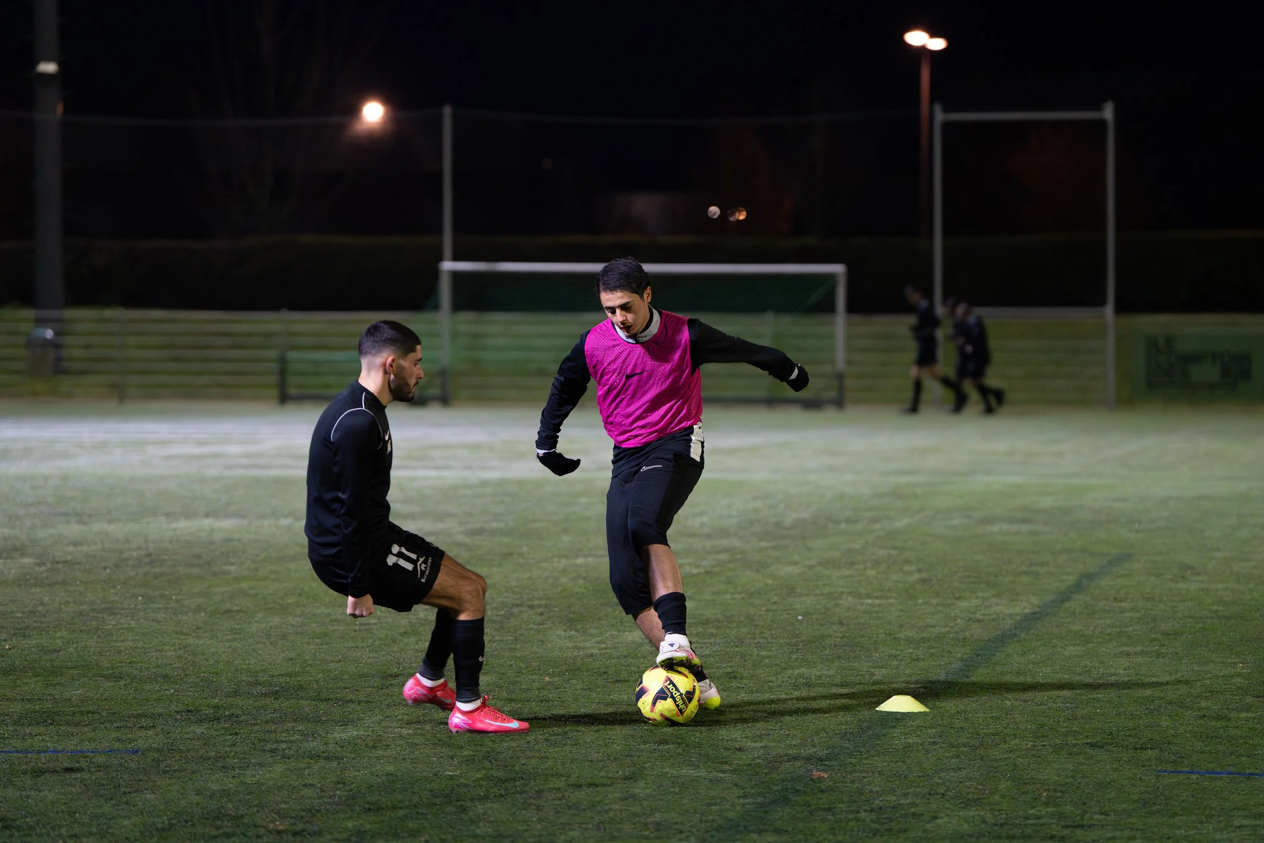 Deux joueurs de football s'entraînent sur un terrain à la tombée de la nuit, avec un ballon et des cônes de marker.