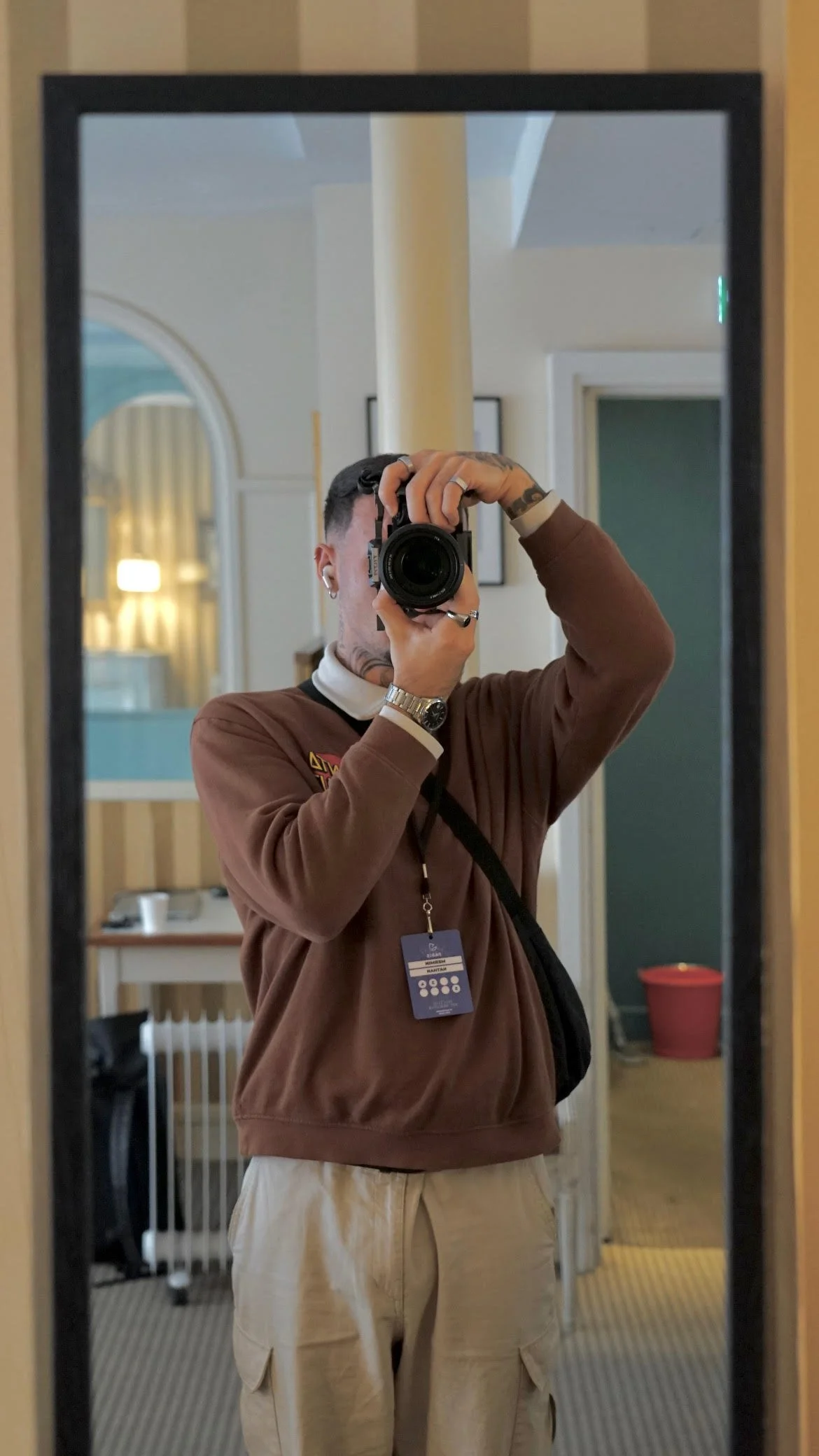 Un homme prenant une photo de lui-même dans un miroir, portant un sweat marron et un pantalon beige, avec des tatouages et des écouteurs, dans une pièce avec des murs en bois et un fond avec une porte verte et un panier rouge.