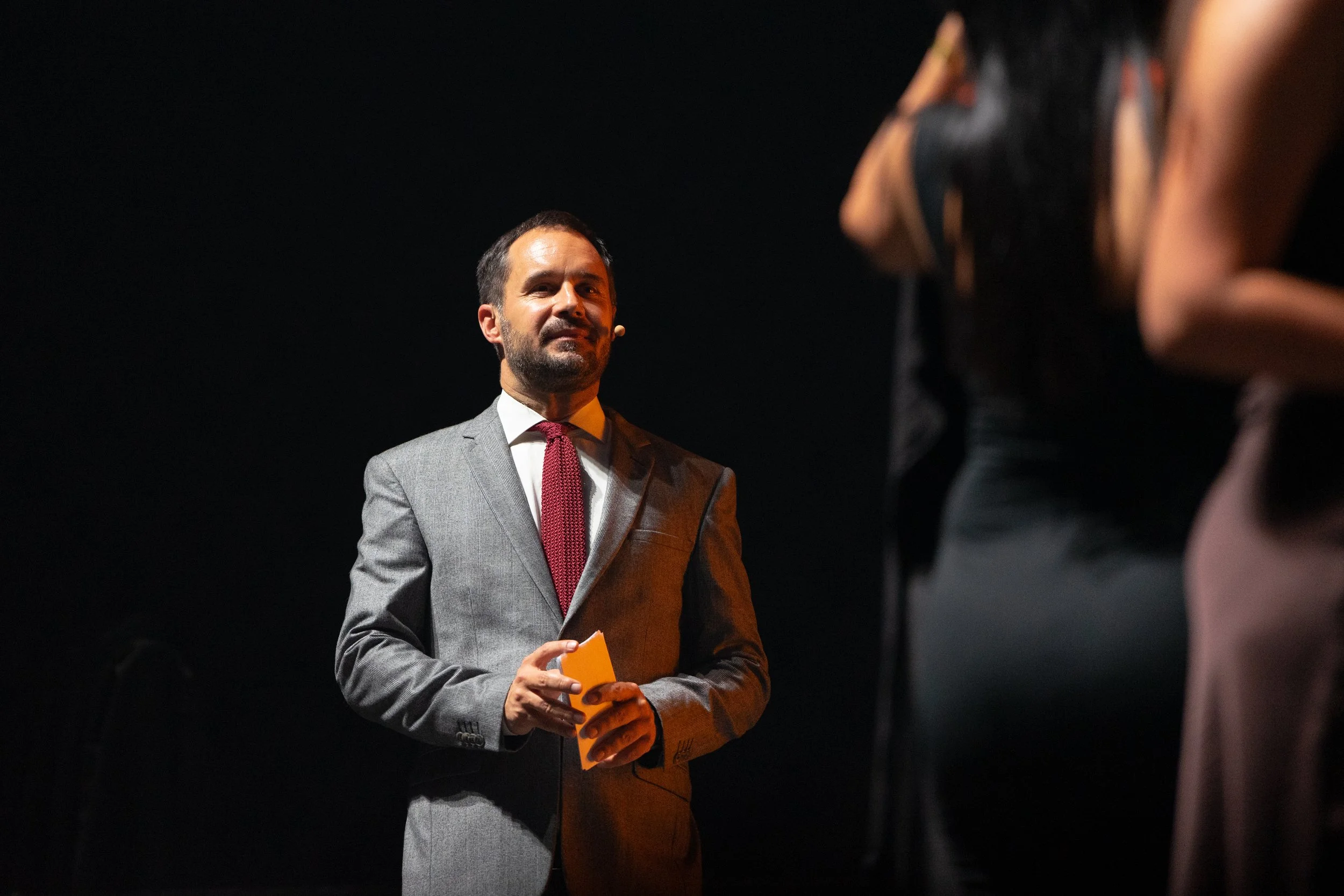 Homme en costume gris avec une cravate rouge, tenant un bloc-notes, sur scène avec un fond noir.