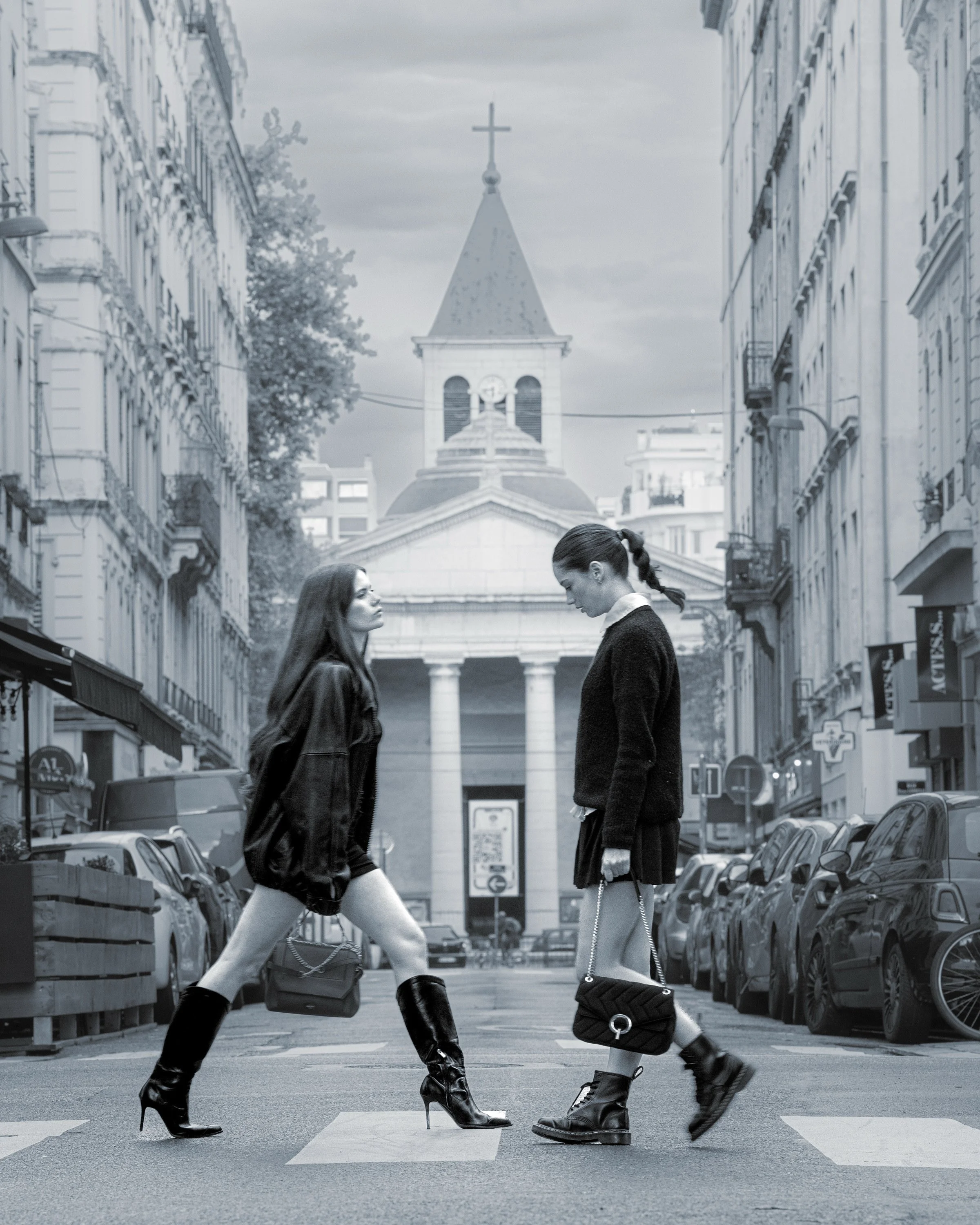 Deux femmes marchent face à face au croisement d'une rue urbaine, avec une église en toile de fond. L'une porte des bottes à talons et une veste en cuir, l'autre des bottines et un petit sac à chaîne. La scène est en noir et blanc.