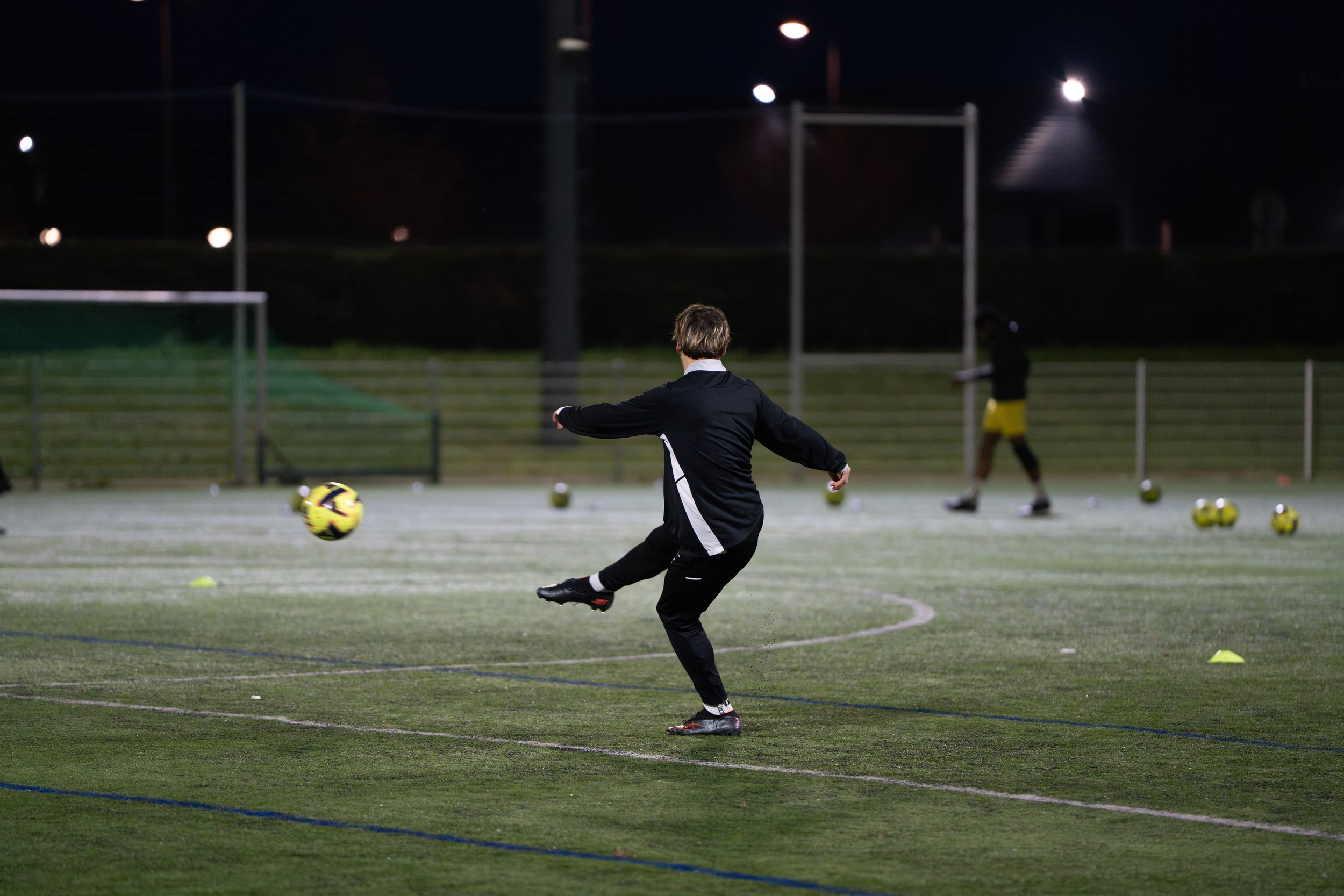 Un entraîneur de football s'entraîne seul sur un terrain de nuit, frappant un ballon avec le pied, avec plusieurs autres ballons éparpillés autour, sous un éclairage nocturne.