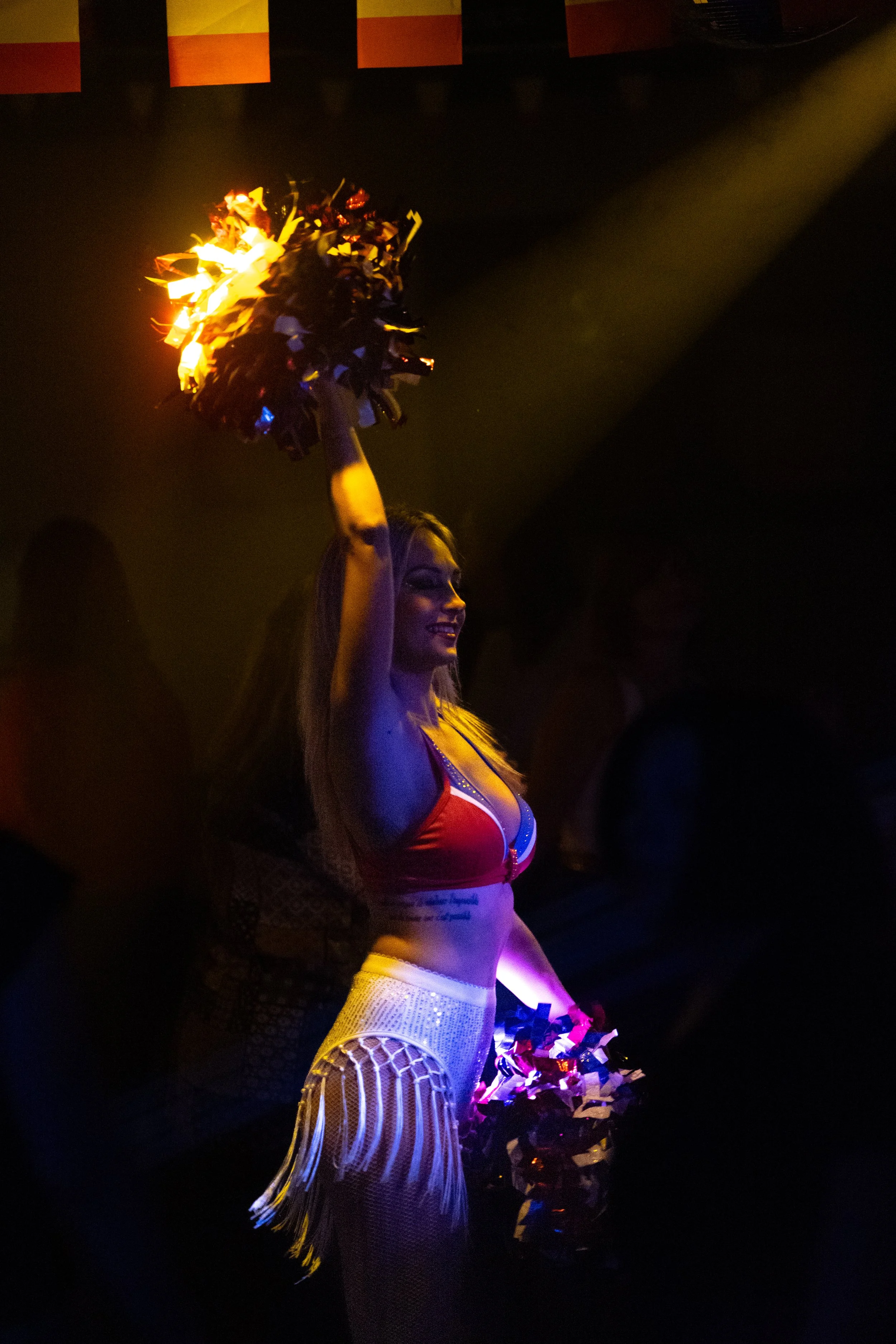 Une femme danseuse en costume coloré, portant des pompons de cheerleading, dans une ambiance sombre éclairée par des lumières chaudes.