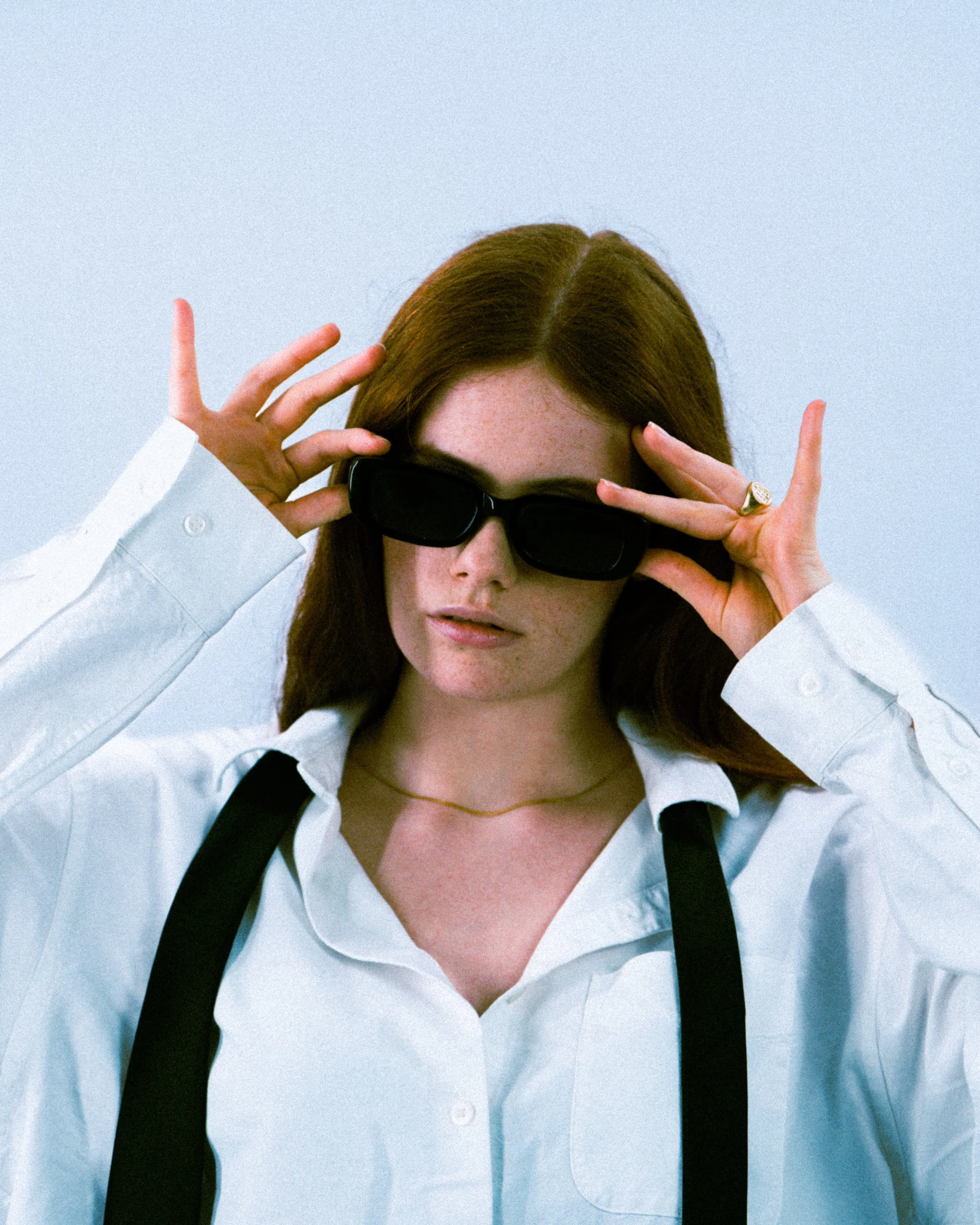 Jeune femme aux cheveux bruns portant des lunettes de soleil noires, une chemise blanche et une cape noire, tenant ses lunettes avec ses mains, regardant vers l'objectif.