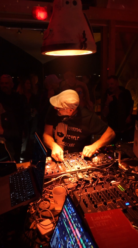 DJ mixant dans un club nocturne avec des lumières rouges, entouré de personnes qui regardent. Il porte un t-shirt noir et un chapeau, avec un ordinateur portable et du matériel de DJ sur la table.