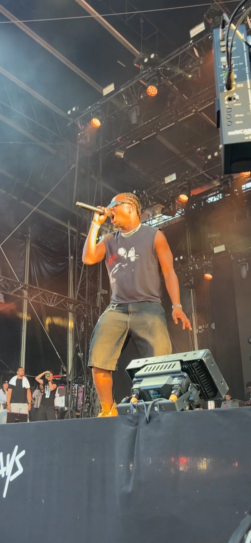 Un artiste performe sur scène lors d'un concert, tenant un microphone, avec un fond de structure métallique et un éclairage orange.