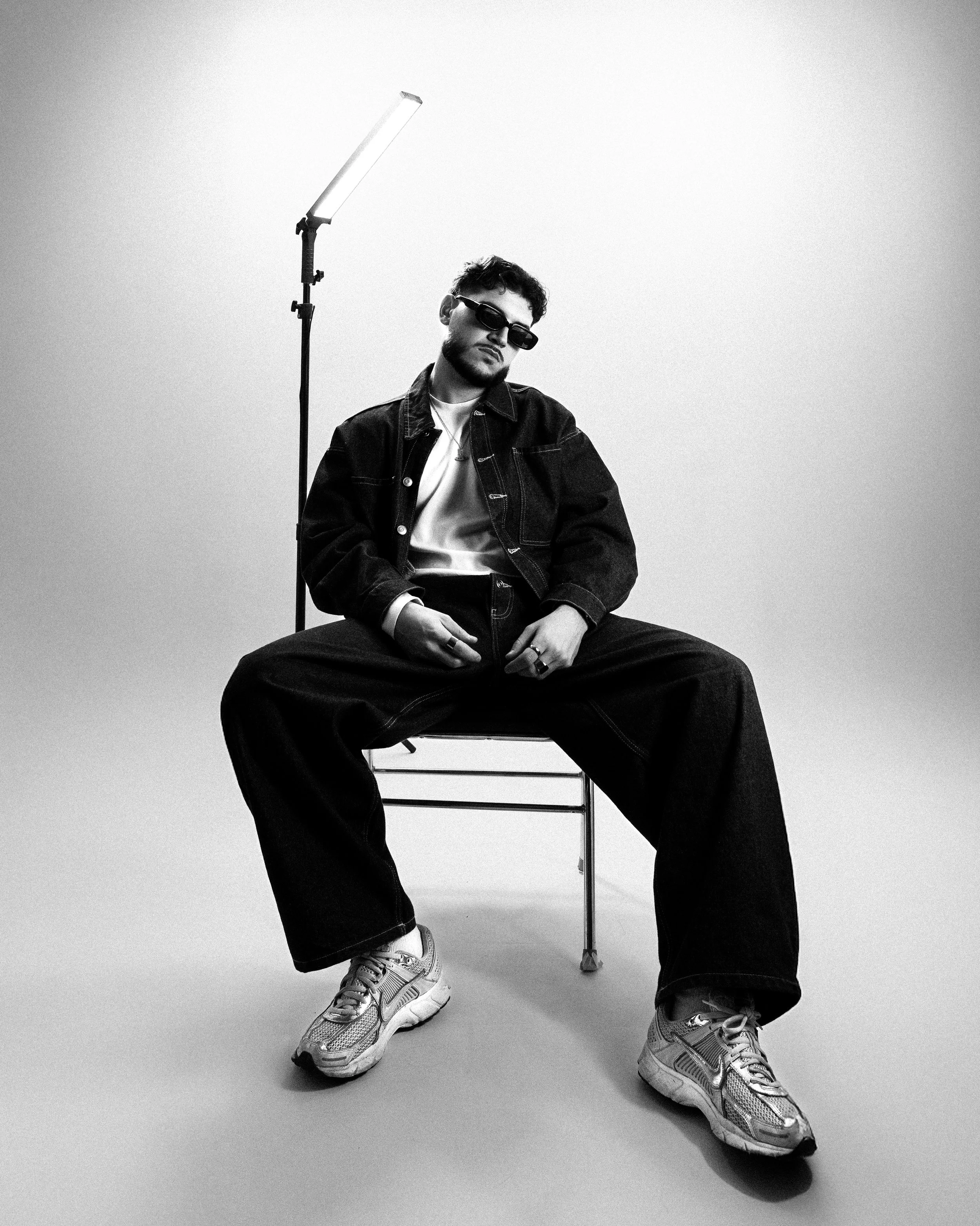 Jeune homme assis sur une chaise, portant des vêtements décontractés, lunettes de soleil, dans un studio avec fond clair, en noir et blanc.