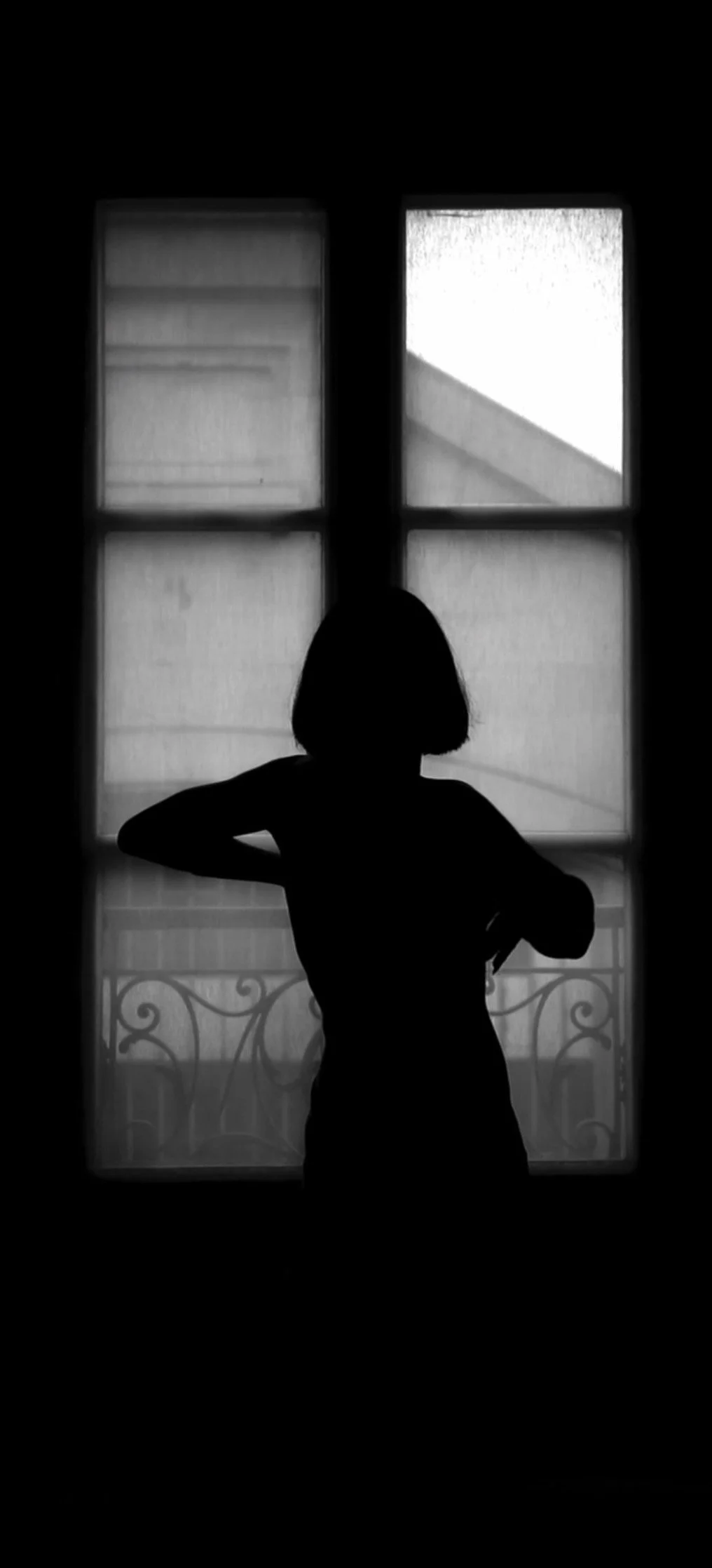 Silhouette d'une femme devant une fenêtre avec des volets transparents, sur un balcon.