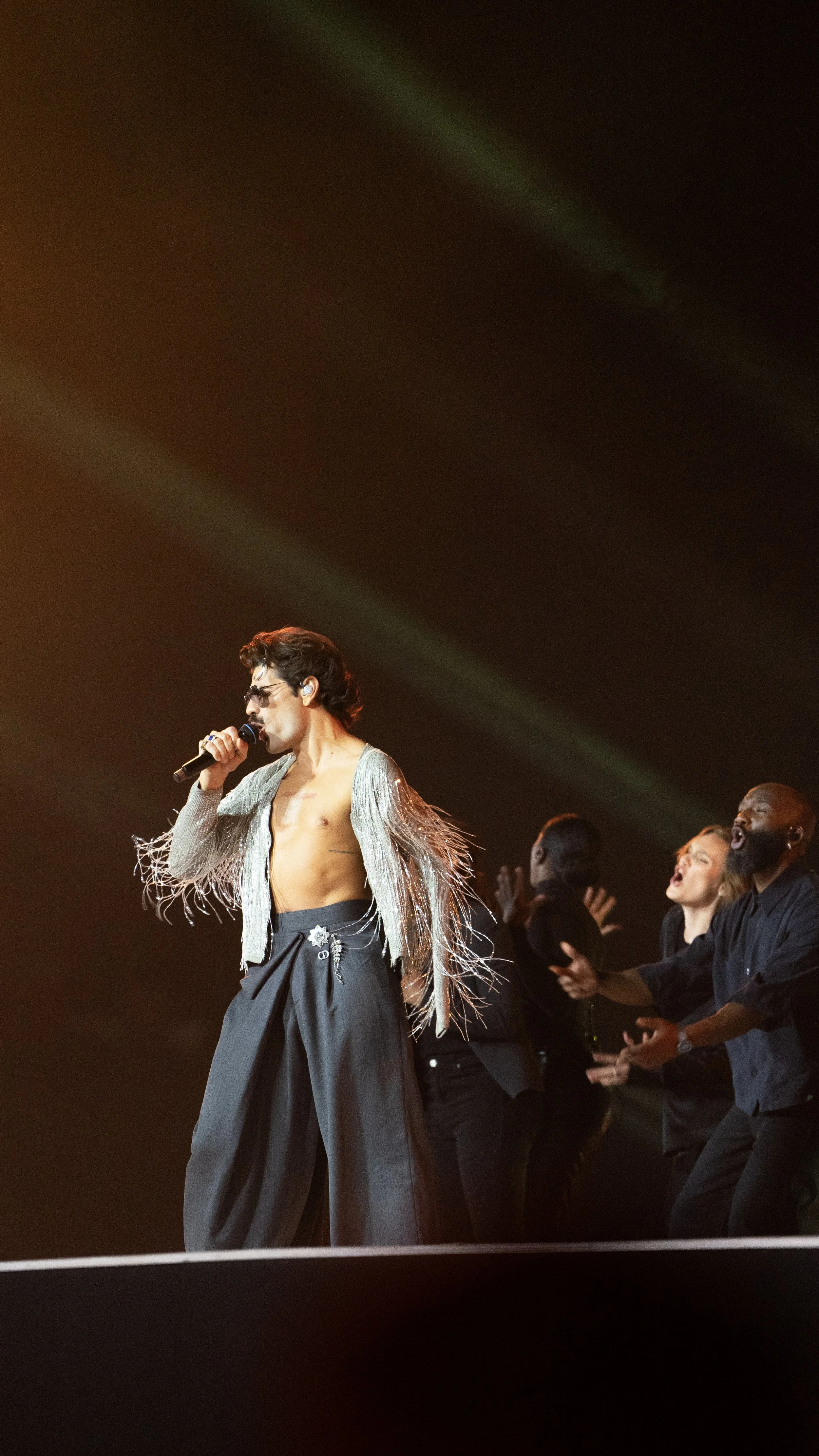 Un homme portant des pantalons noirs et un haut en paillettes, tenant un micro, en train de chanter sur scène, avec un groupe de choristes derrière lui.