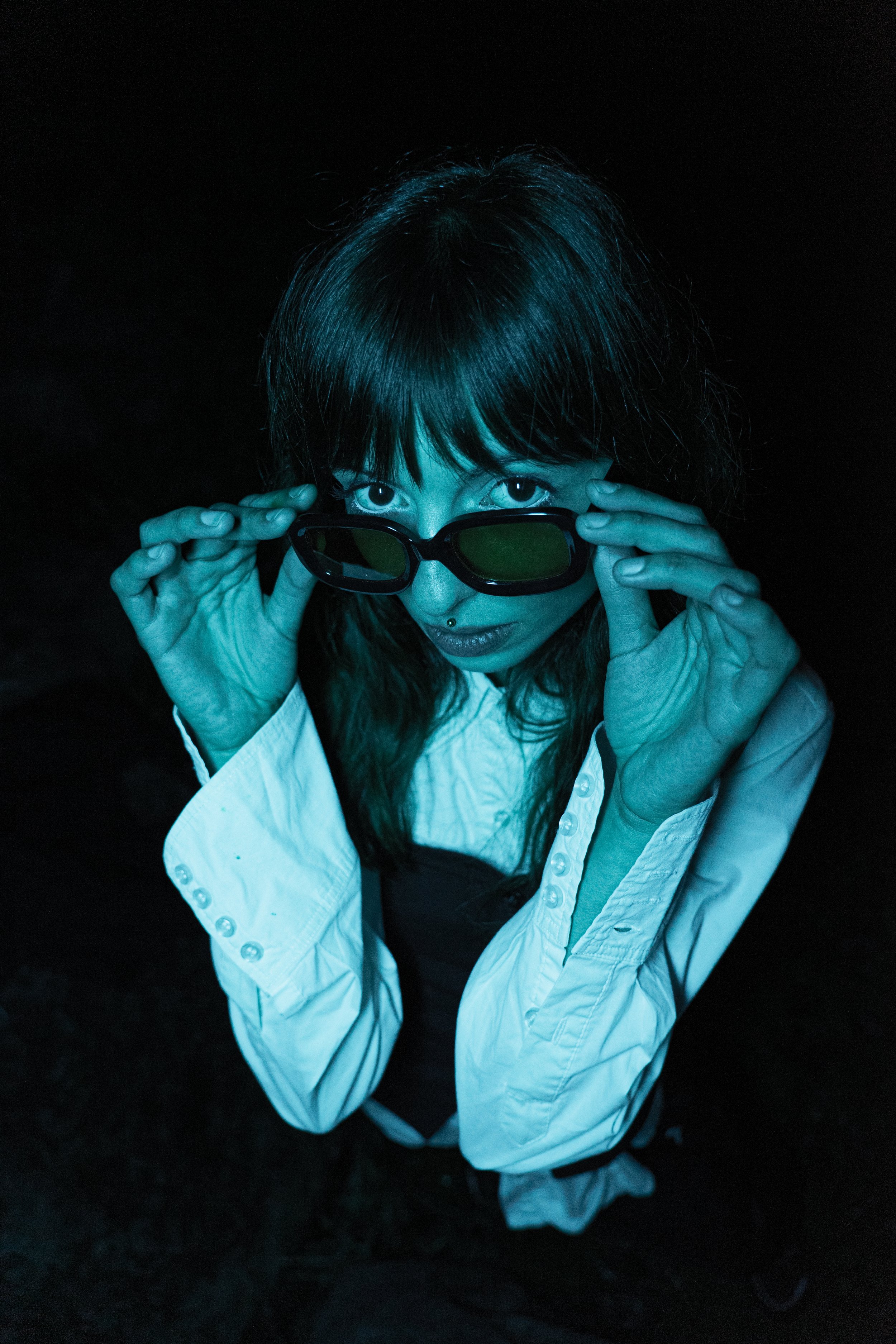 Une femme regarde au-dessus de ses lunettes de soleil, habillée en blanc et noir, dans un environnement sombre.