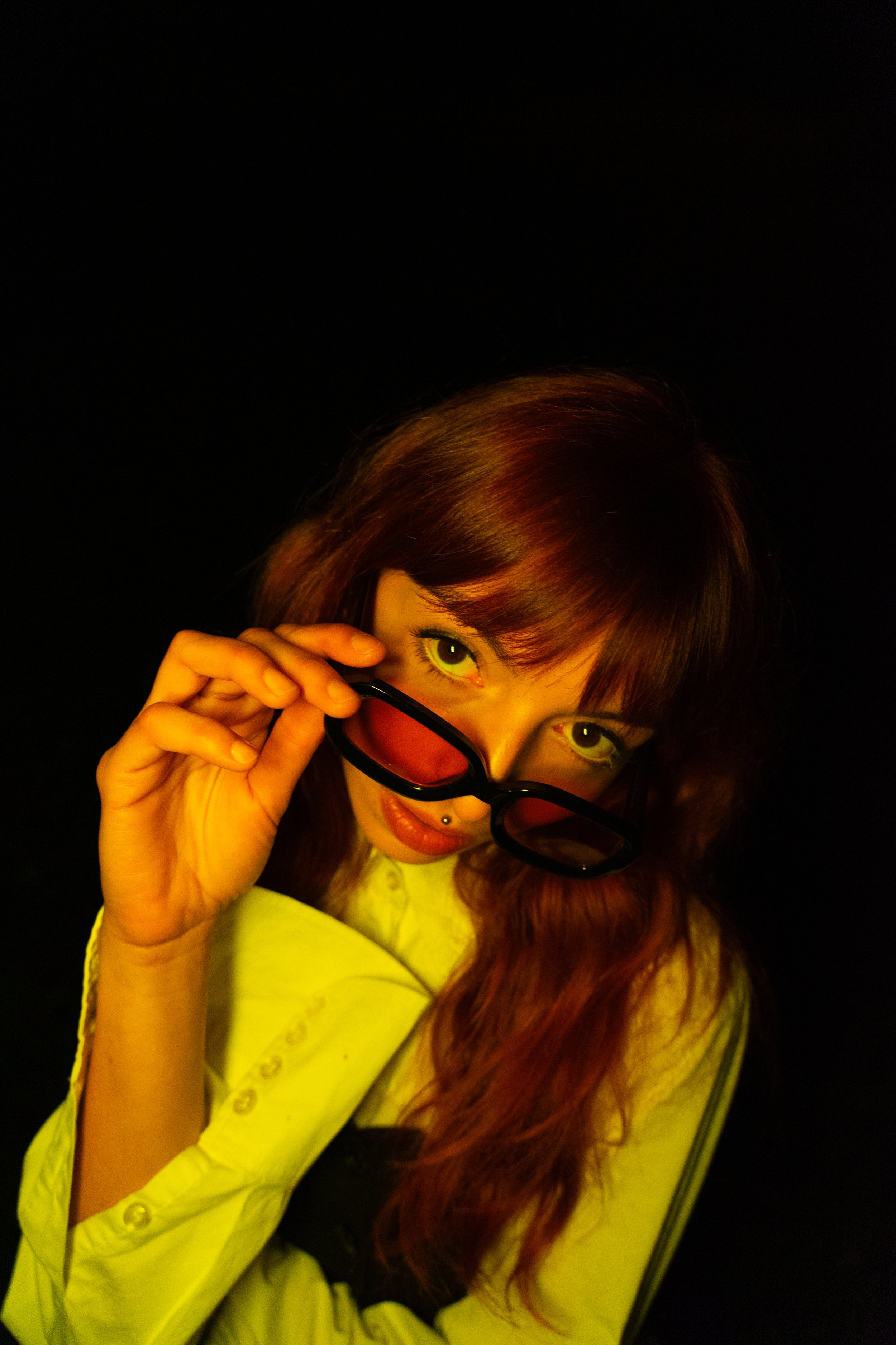 Femme avec longs cheveux bruns, portant des lunettes de soleil noires, regardant par-dessus en les tenant de sa main, pull jaune clair, fond noir.