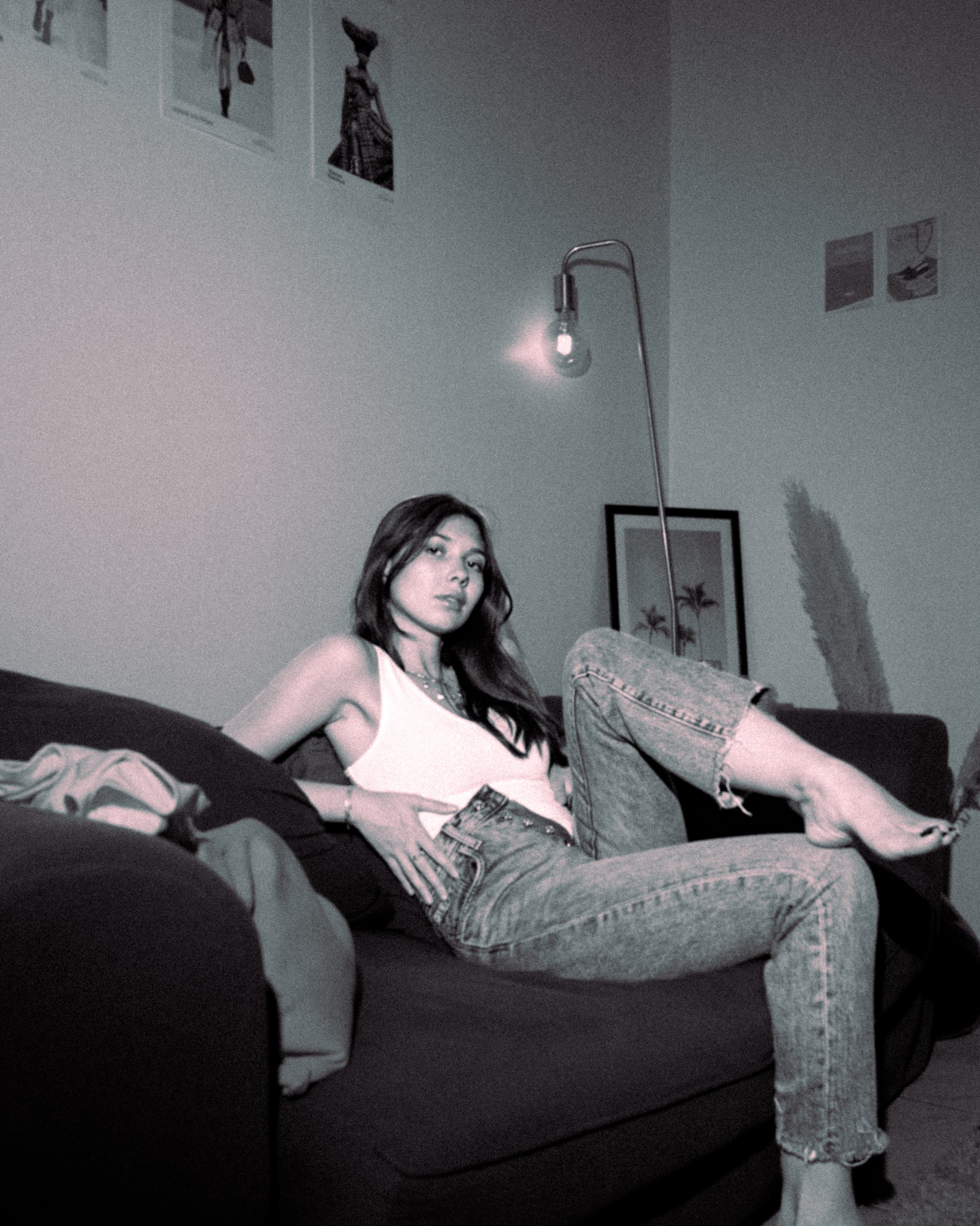 Une jeune femme assise sur un canapé, portant un débardeur blanc et un jean déchiré, dans une pièce avec des décorations murales et un lampadaire suspendu.