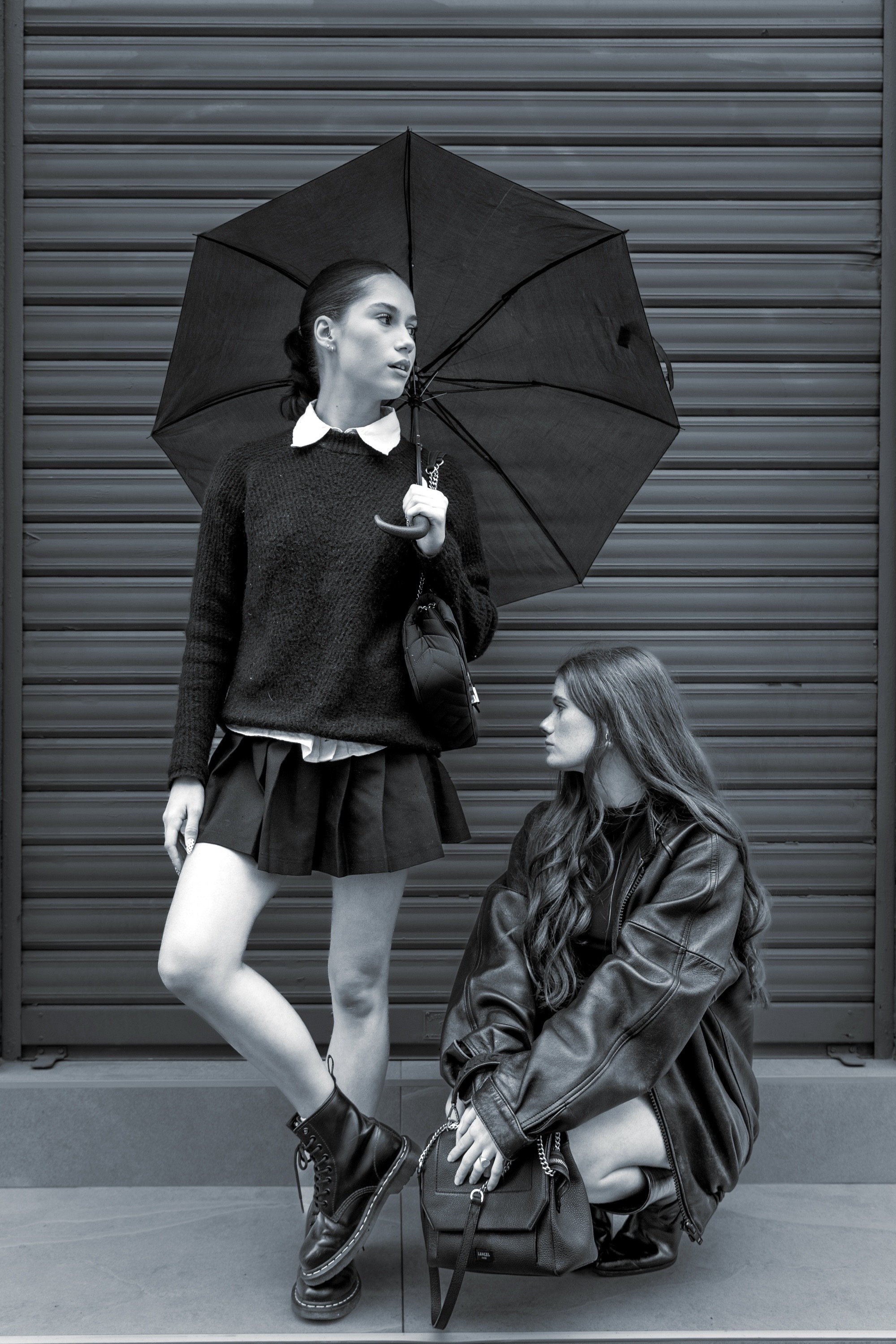 Deux jeunes femmes avec des vêtements modernes, une tenant un parapluie noire, devant une porte de garage en métal, en noir et blanc.