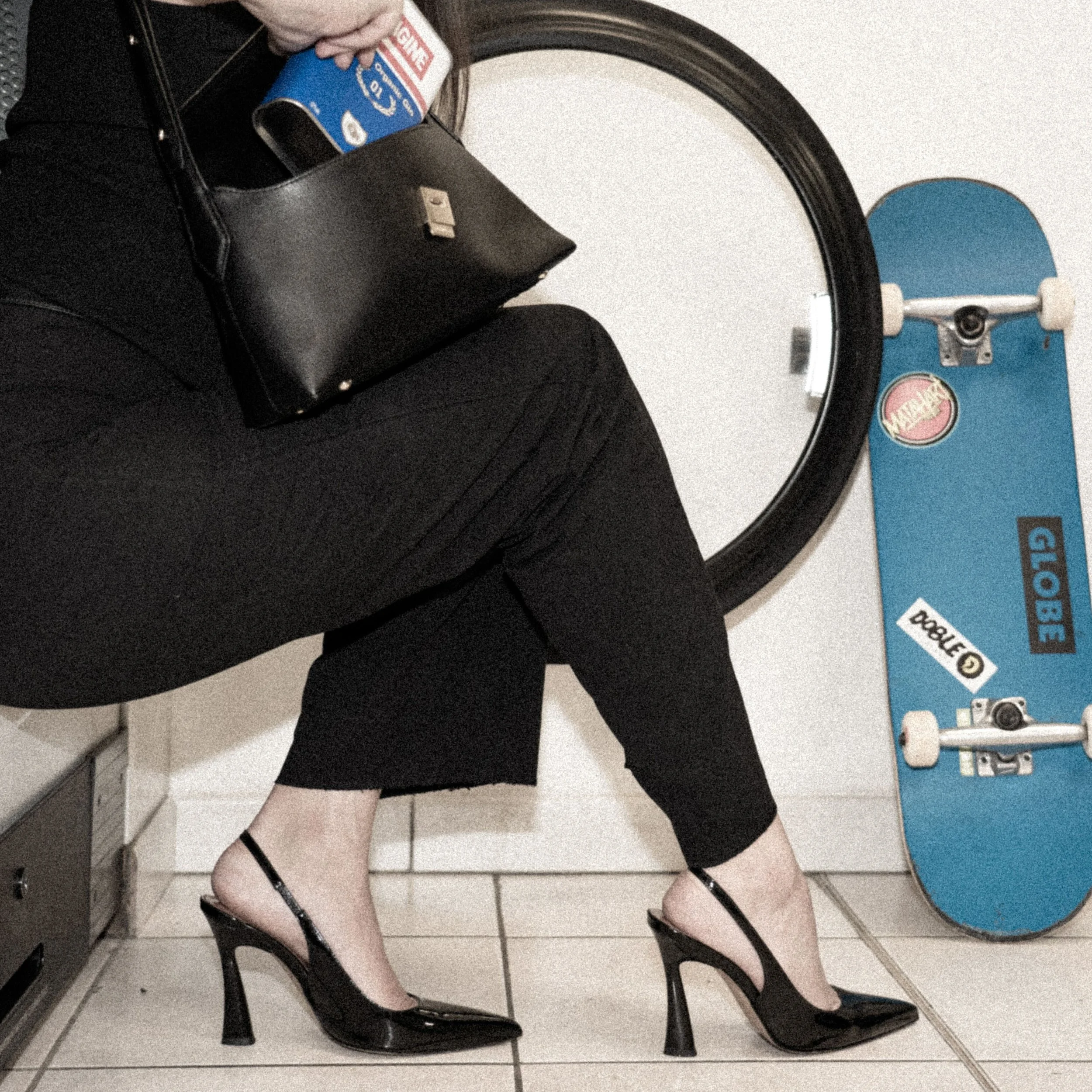 Une personne habillée en noir, portant des talons hauts noirs et une jupe ou pantalon noir, assise en position décontractée avec une valise noire à côté, dans un lieu probablement une salle de bain ou un espace public, avec un skate bleu et un miroir