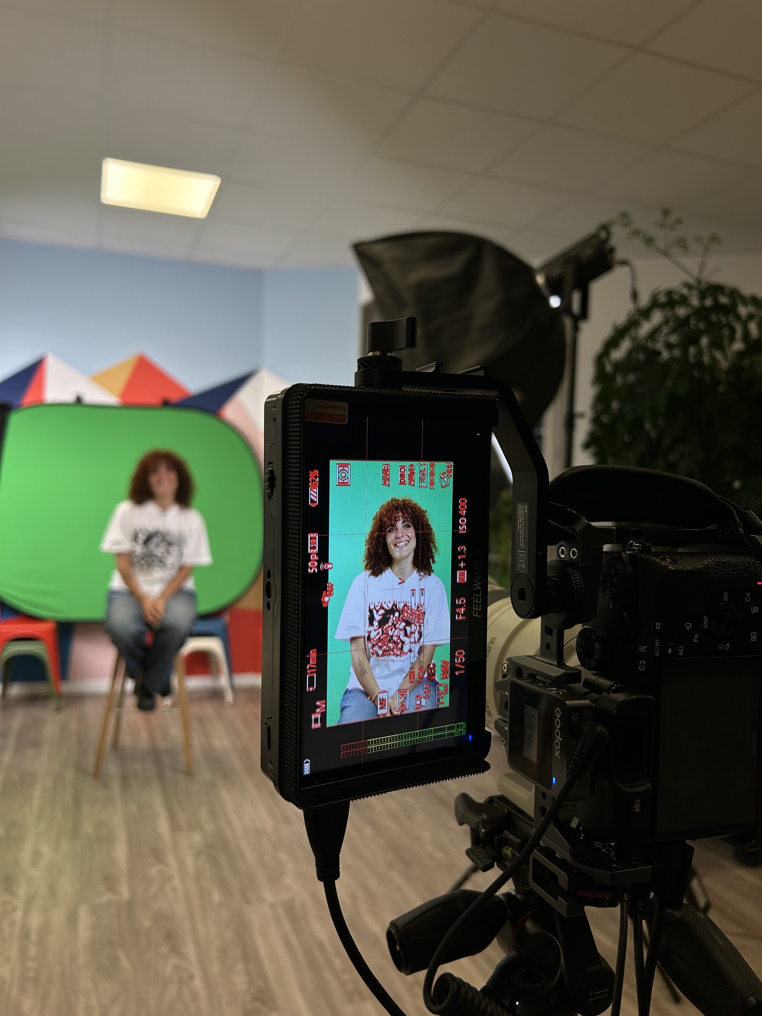 Une personne coiffée de boucles bouclées, portant un t-shirt gris avec un motif rouge, assise sur une chaise devant un fond vert, dans un studio avec éclairage professionnel.