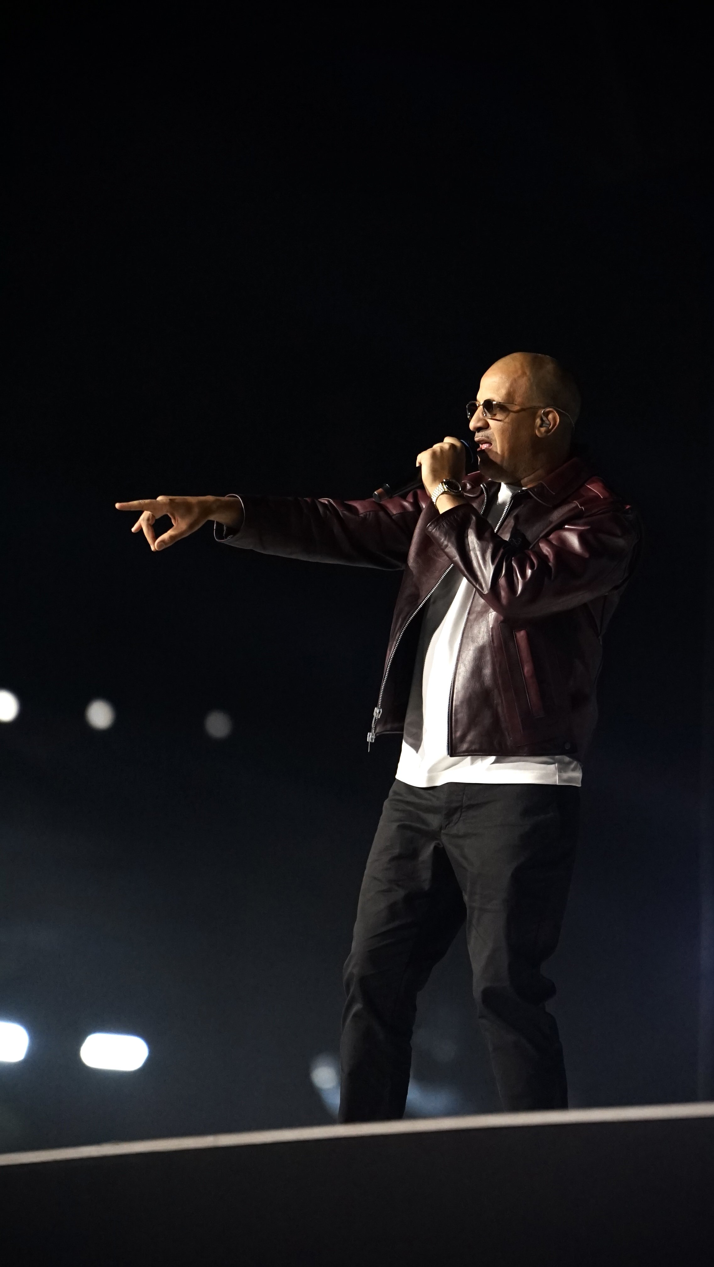 Homme chantant avec un micro, portant des lunettes de soleil, veste en cuir bordeaux, bas dans une scène sombre avec des lumières en arrière-plan.