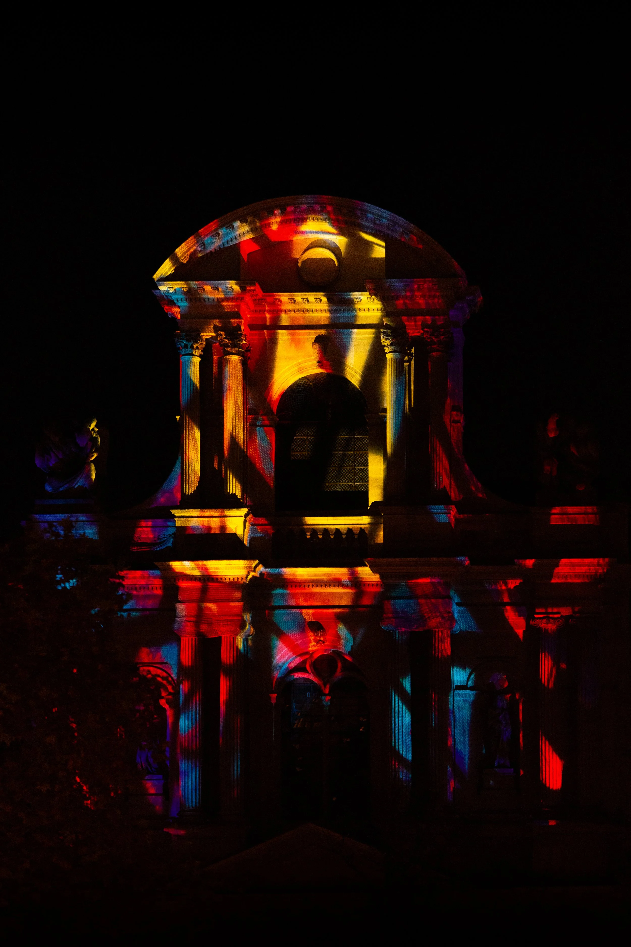 Projection de lumières colorées sur un bâtiment historique en pierre, avec colonnes et statues, à la nuit.