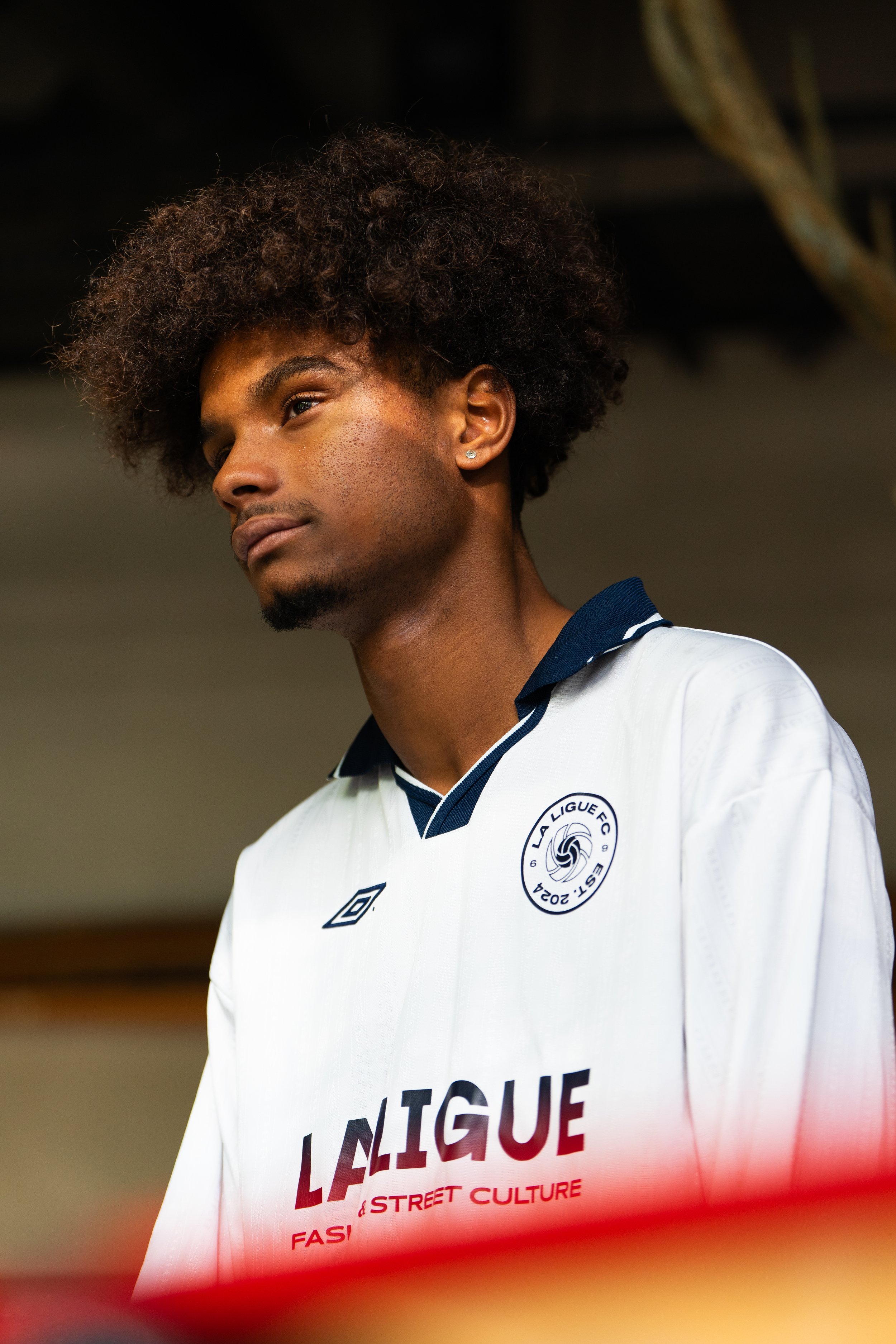 Un homme portant un maillot de football blanc avec le logo de La Ligue FC, regardant pensivement, dans un environnement intérieur.