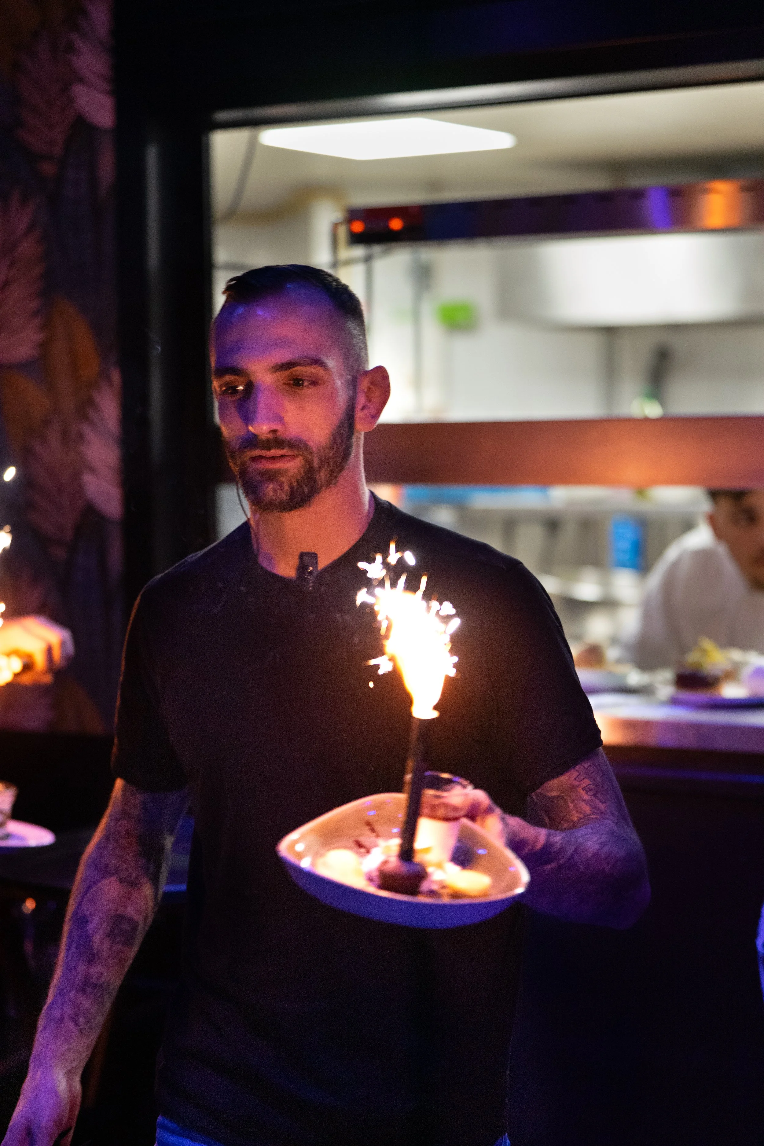 Un homme avec une barbe tenant une assiette avec un gâteau et une bougie flashant, dans une cuisine ou un restaurant.