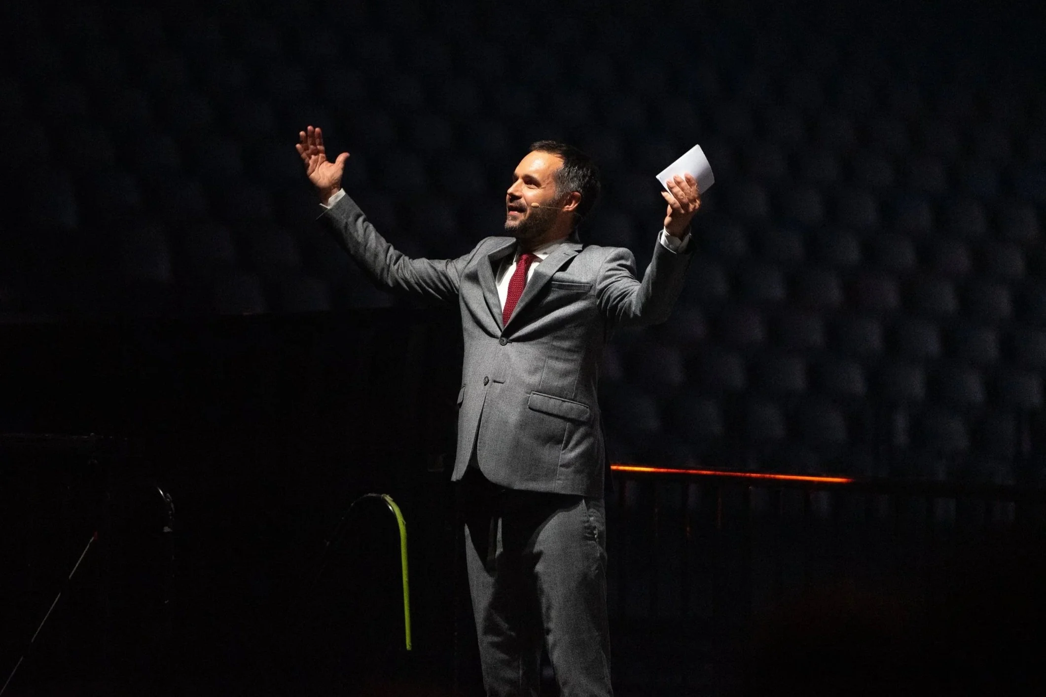 Homme en costume gris, levant les bras, parlant ou présentant quelque chose, dans un environnement sombre.