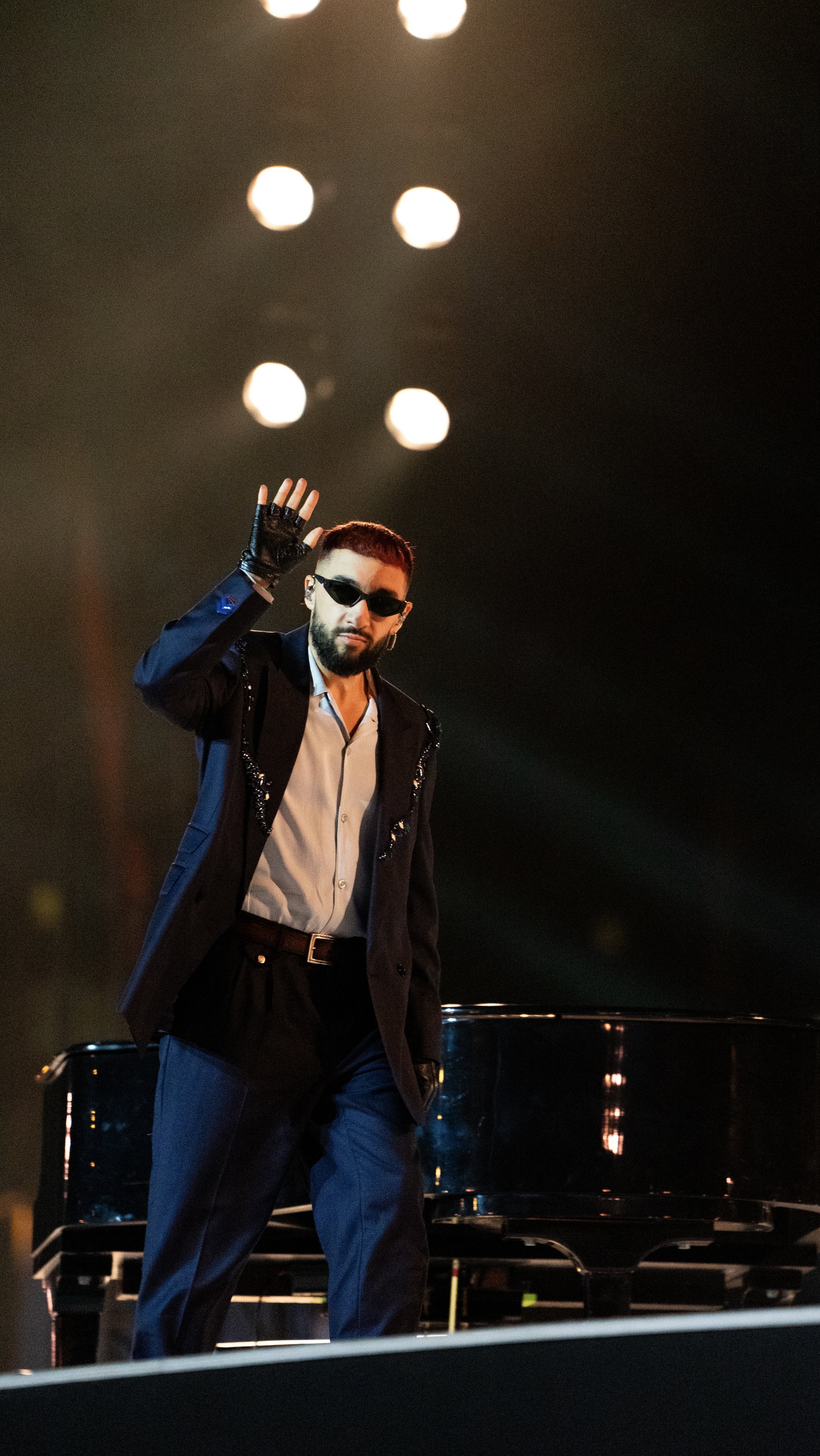 Un homme habillé en noir avec des lunettes de soleil, saluant sur scène près d'un piano noir, en arrière-plan des lumières blanches