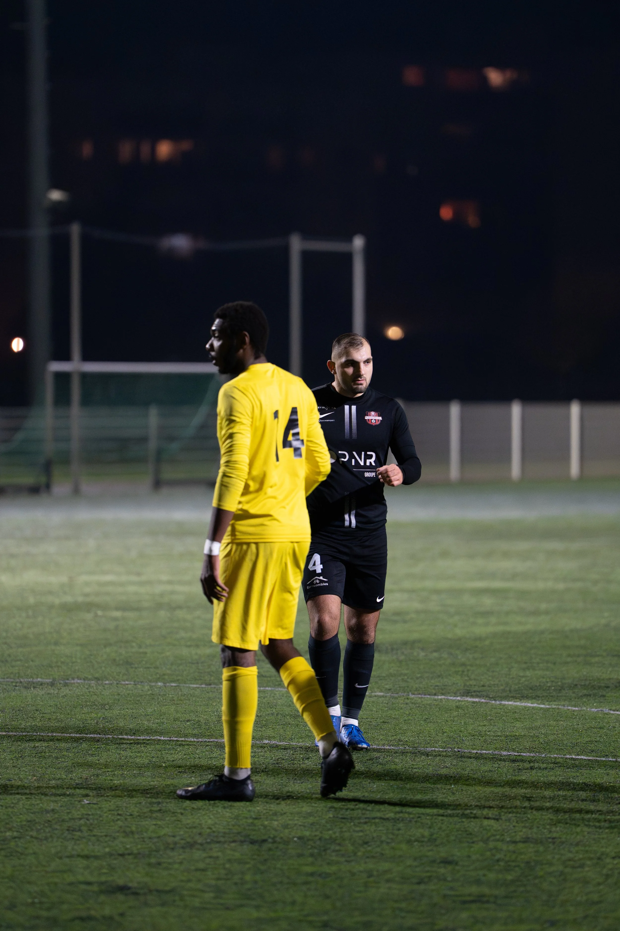 Deux joueurs de football en tenue de match sur un terrain de nuit, un en jaune et l'autre en noir, échangeant probablement des mots ou des gestes.