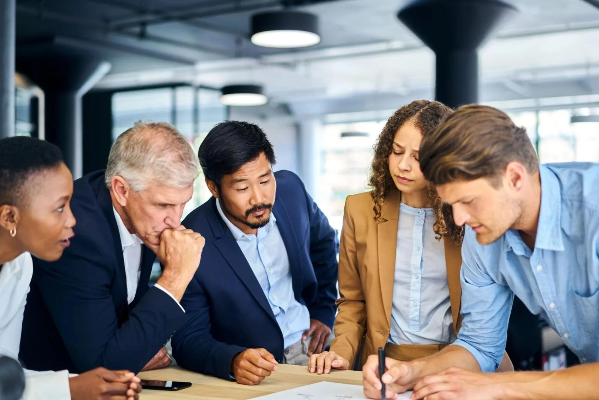 A diverse group of six professionals, three women and three men, gathered around a table in a modern office, engaged in a serious discussion or brainstorming session.