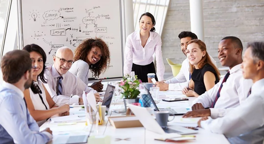 A diverse group of professionals in a business meeting in a bright, modern conference room. They are sitting around a white table with laptops, papers, and coffee cups, smiling and engaging. A woman stands at the whiteboard with handwritten notes and diagrams.