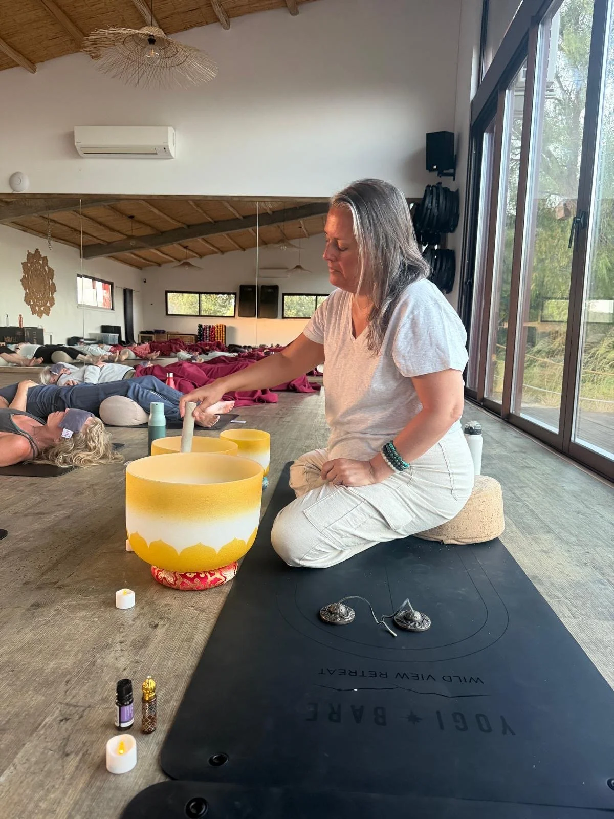 A woman sitting on a cushion in a yoga studio, leading a group through a sound healing session with singing bowls. Participants are lying on mats in the background during the session.