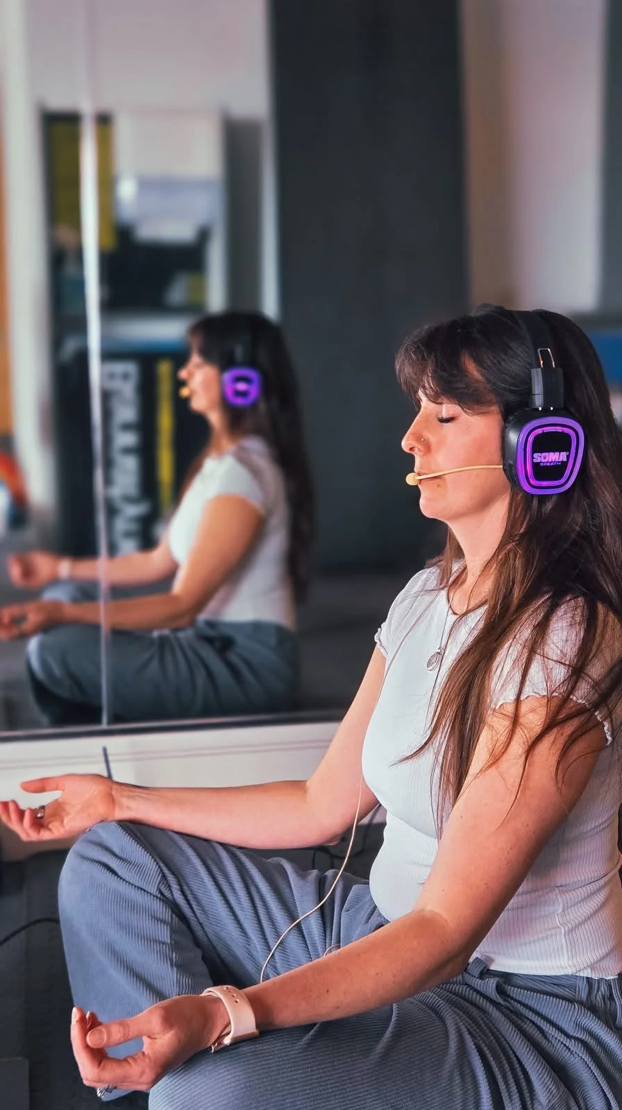 A woman sitting cross-legged with eyes closed, wearing headphones and a microphone, meditating in a fitness or wellness setting.