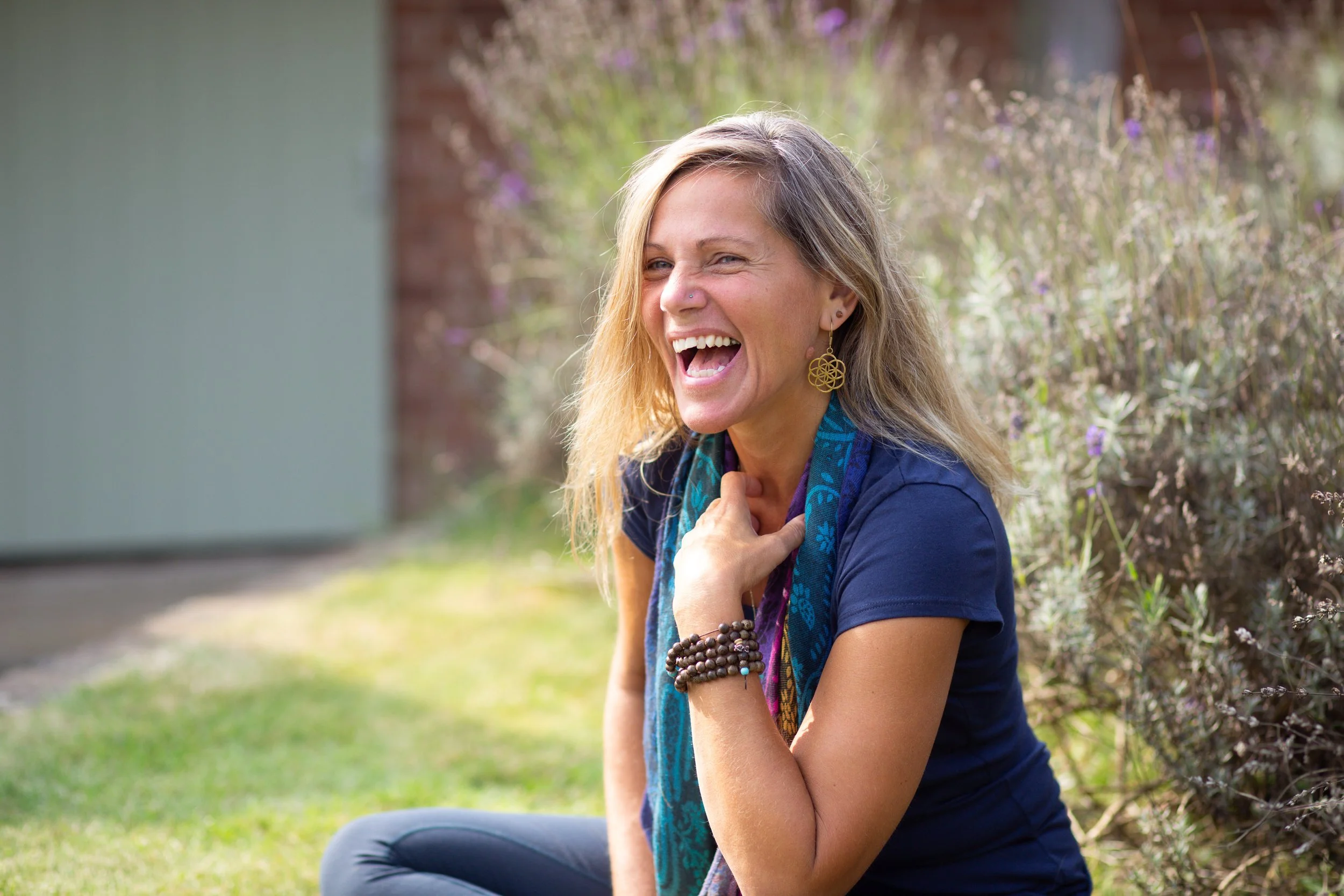 Woman laughing outside, sitting on the grass, wearing a blue shirt, colorful scarf, and gold earrings, with purple flowers in the background.