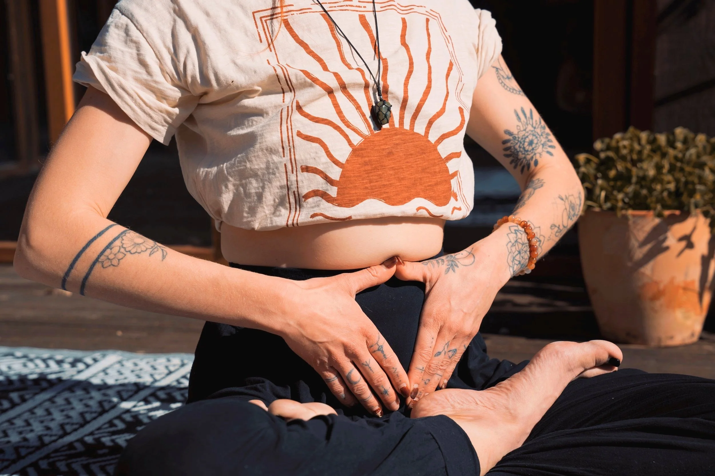 Person sitting cross-legged outdoors in a yoga pose, wearing top with a sun design and holding hands over the abdomen, on the decking of SunFyr barns.