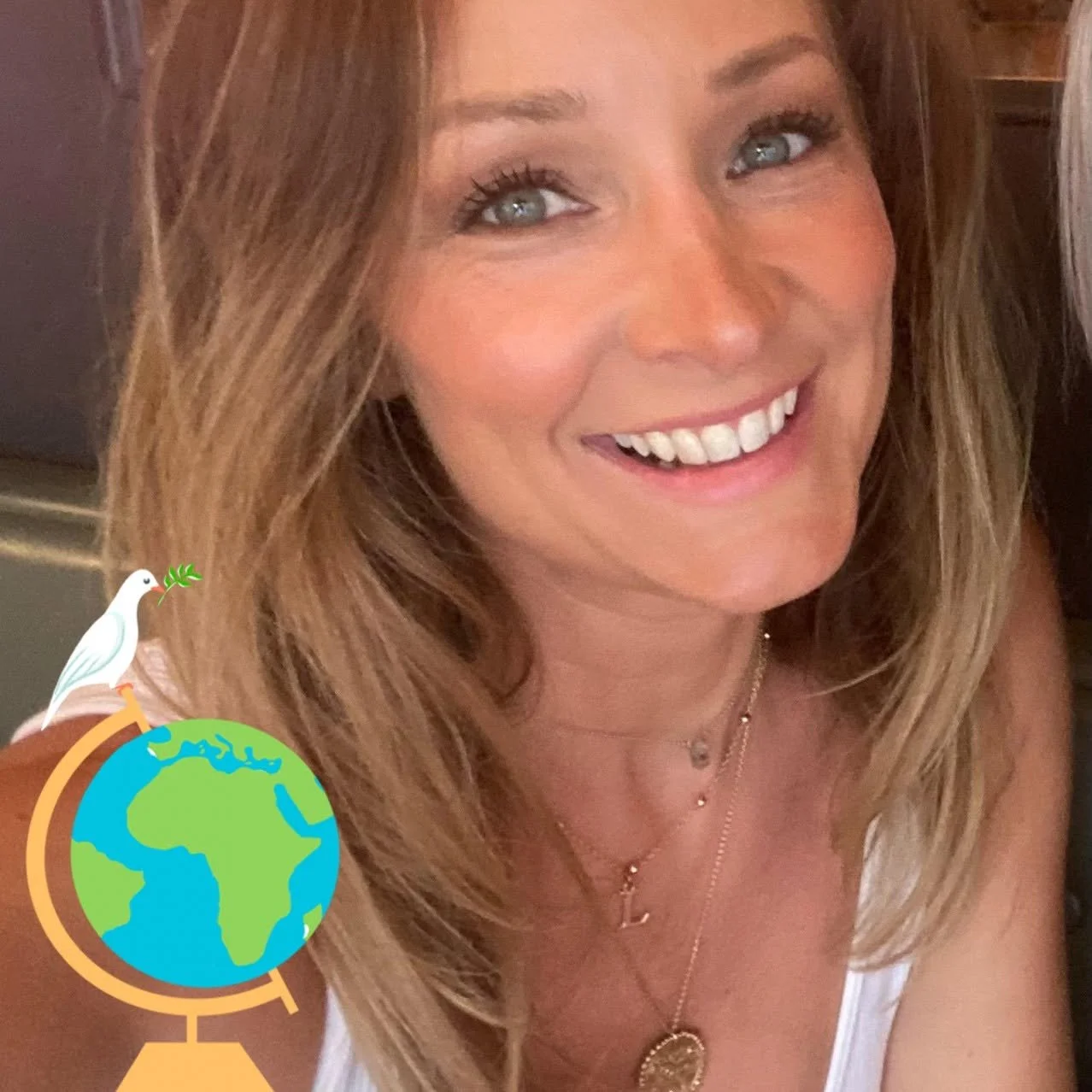 Close-up of a smiling woman with blonde hair, wearing jewelry, and a white top, with a digital Earth and dove sticker in the bottom corner.