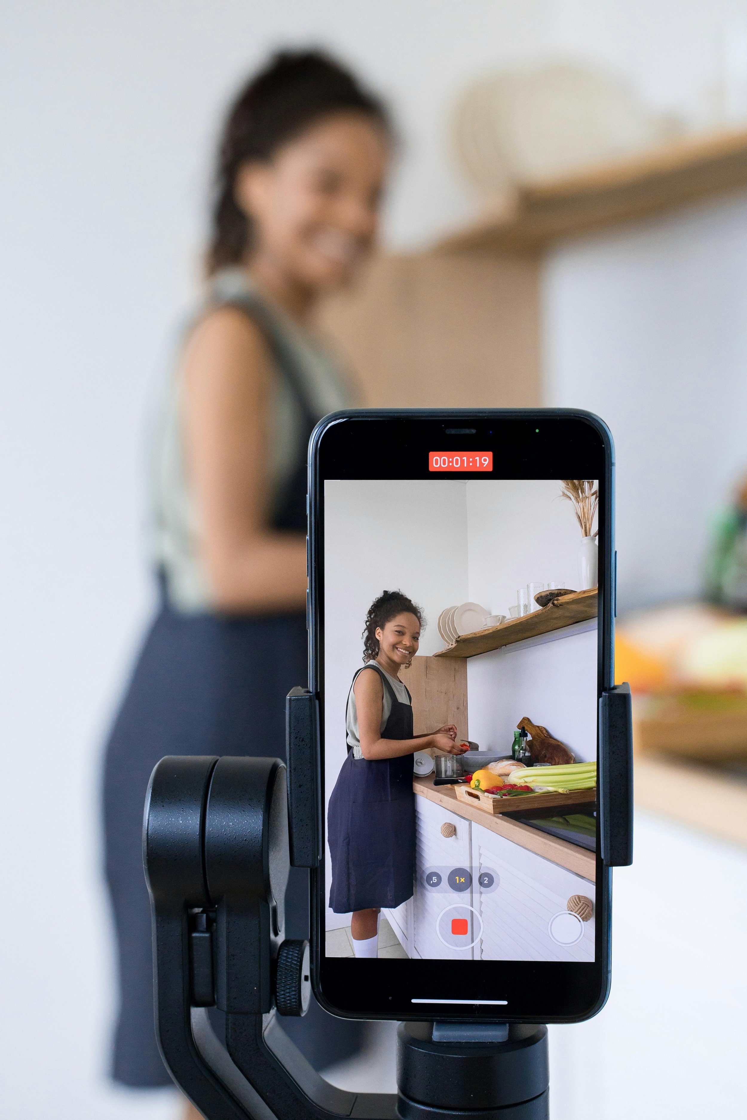 A young woman is recording a video of herself in the kitchen, smiling and preparing food behind a camera on a tripod, with a kitchen shelf and various kitchen items visible in the background.