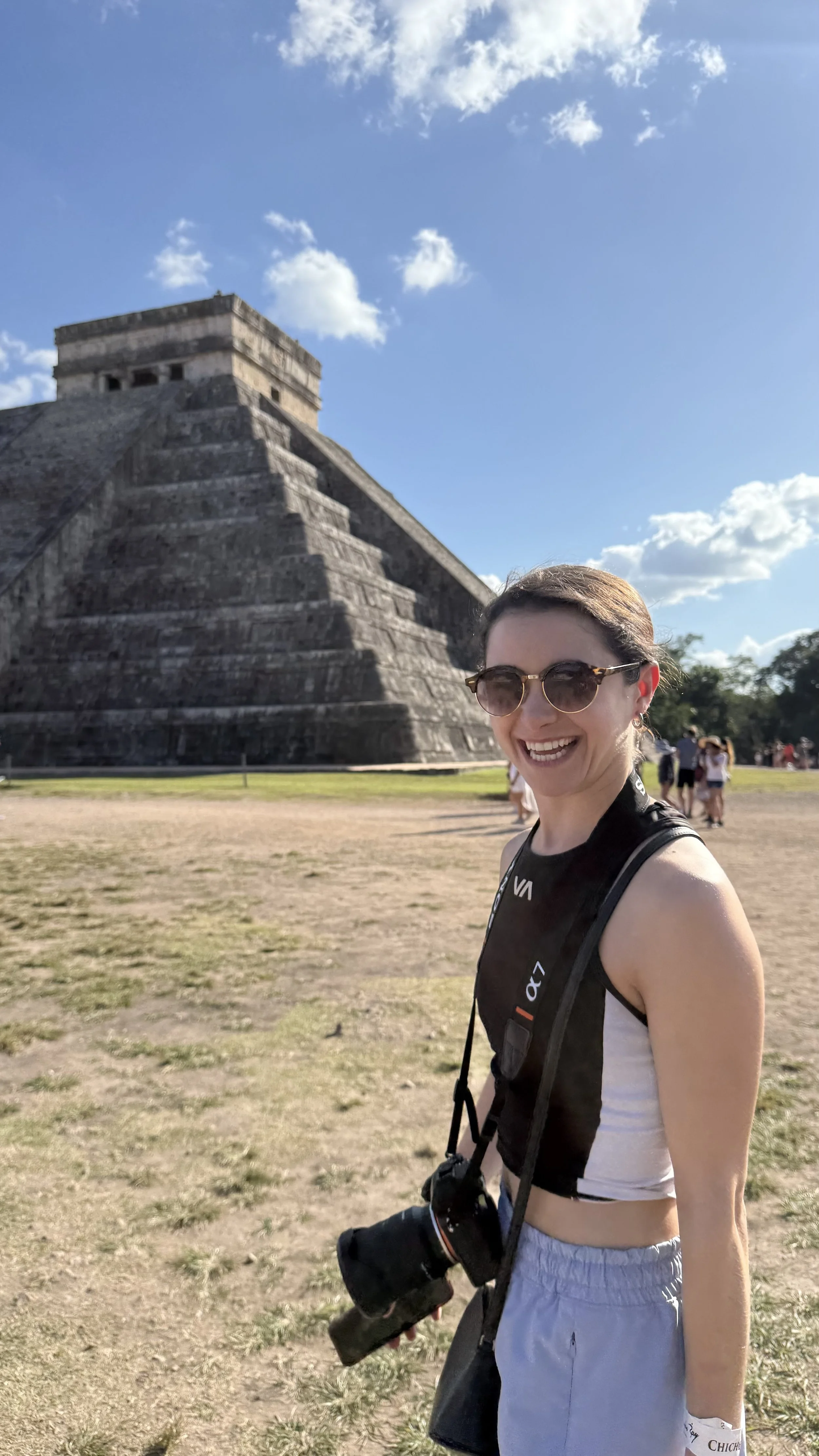 A young woman with sunglasses, a camera hanging around her neck, and casual clothing standing in front of the ancient Mayan pyramid El Castillo at Chichen Itza in Mexico, smiling at the camera.