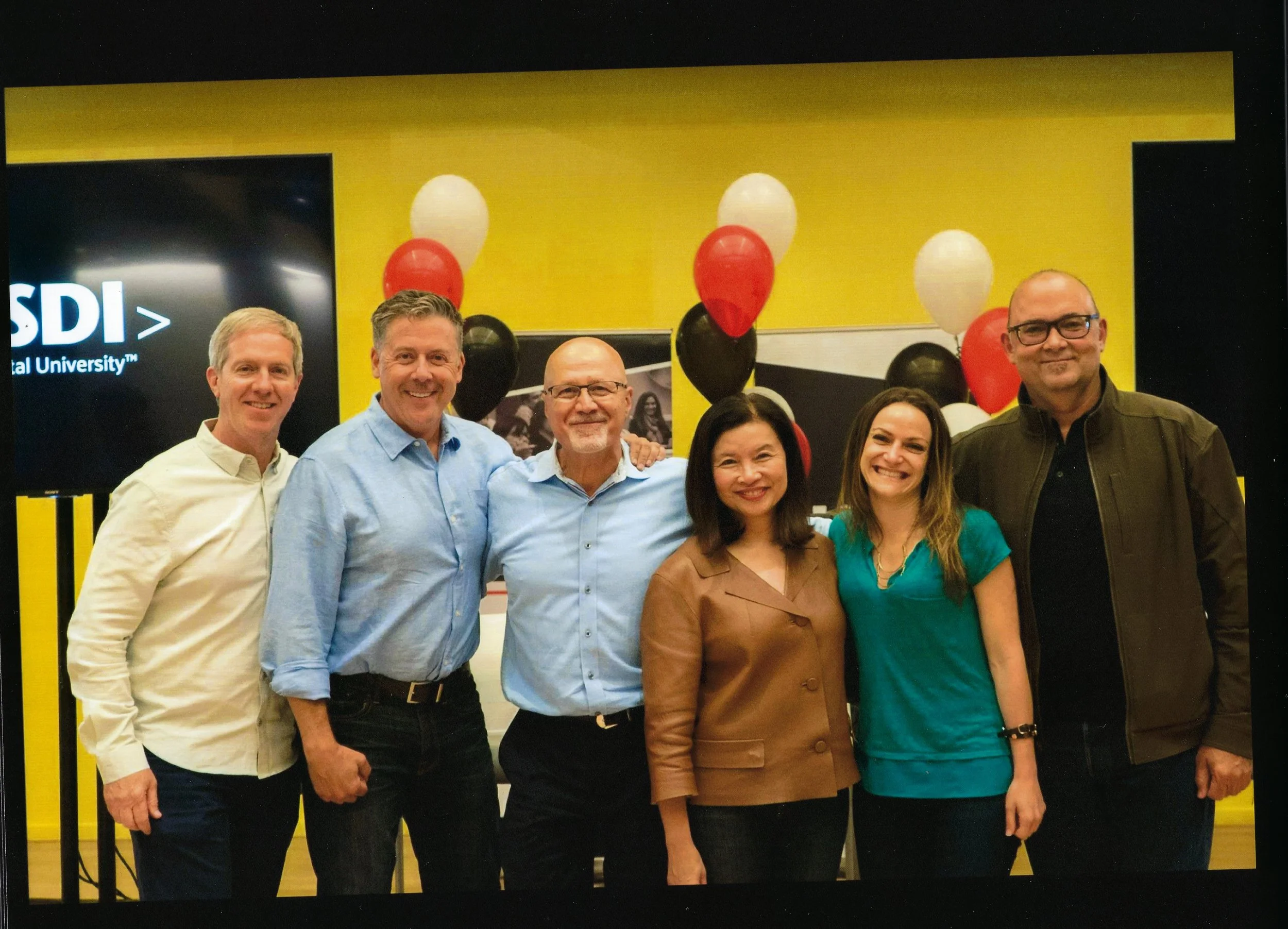 A group of seven smiling adults standing in front of a yellow wall with black and white balloons and a black banner with white text that says "SDI Digital University".