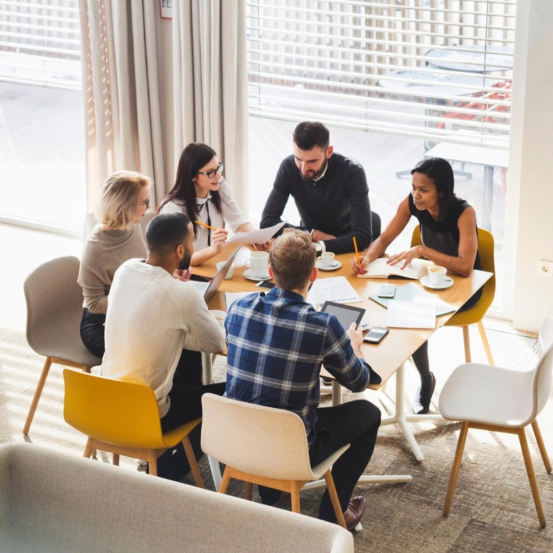 A group of diverse people sitting around a table in a bright room with large windows having blinds, engaged in a discussion, with notebooks, cups, and electronic devices on the table.