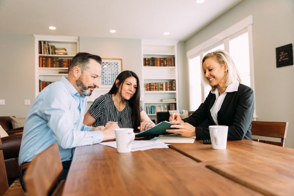 Three people sitting at a wooden table, discussing documents, with bookshelves in the background and sunlight coming through windows.