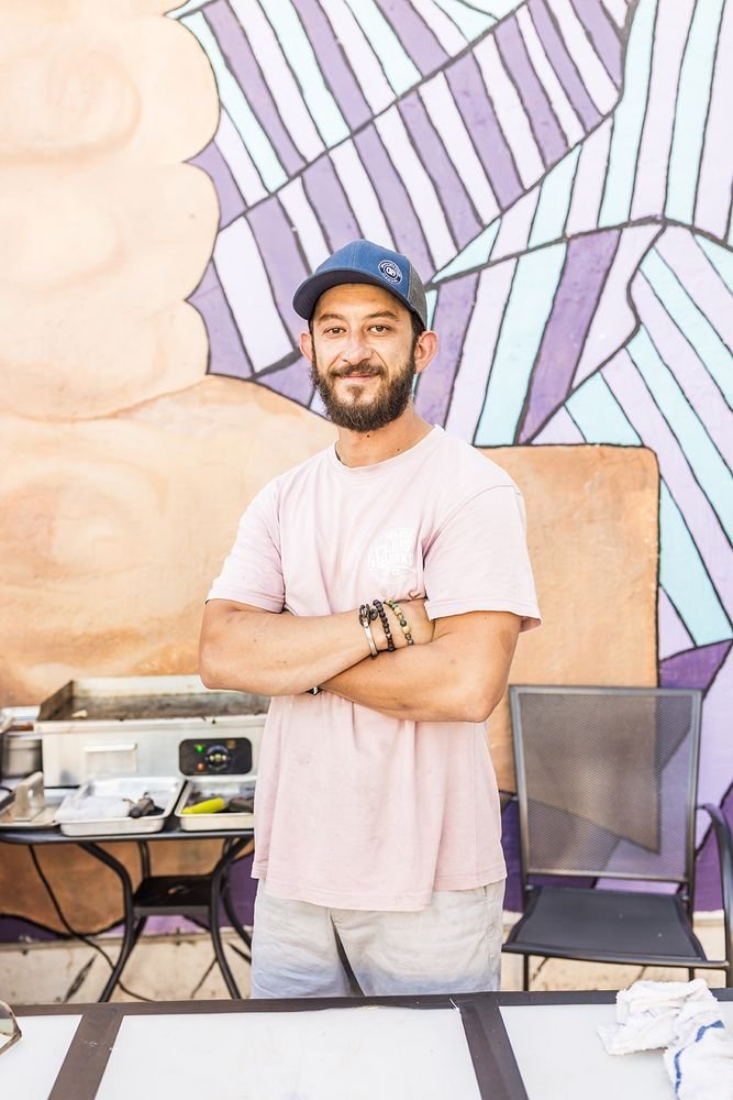 Man chef efe onoglu with arms crossed wearing a cap and pink shirt, standing in front of a colorful mural with geometric patterns. He is smiling, and there is outdoor cooking equipment on a table beside him.