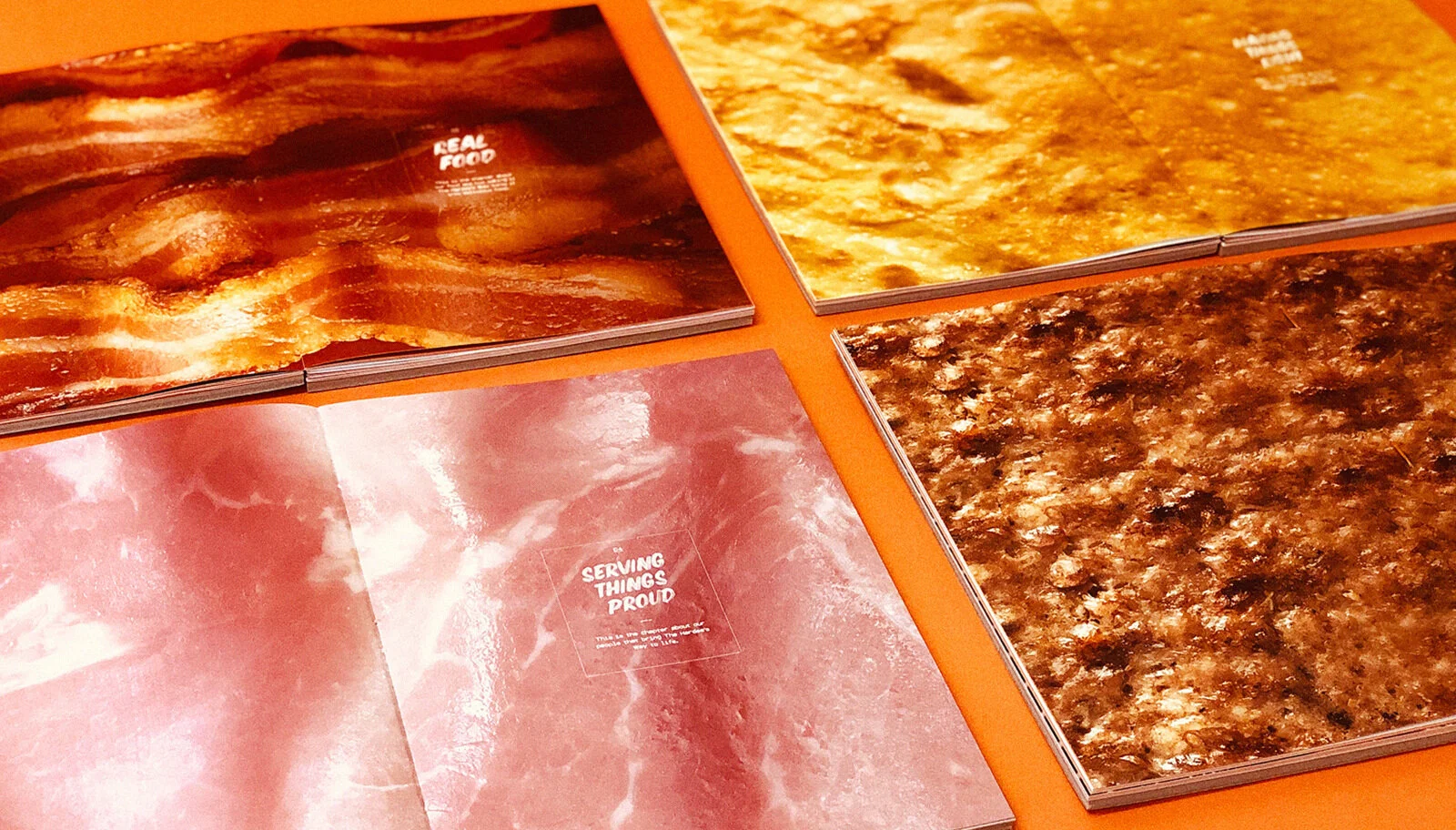 Four open magazines with food imagery on an orange surface. Top left magazine shows cooked bacon, top right magazine shows a cooked dish, bottom left magazine shows a pinkish surface with text, and bottom right magazine shows a cooked dish with brown