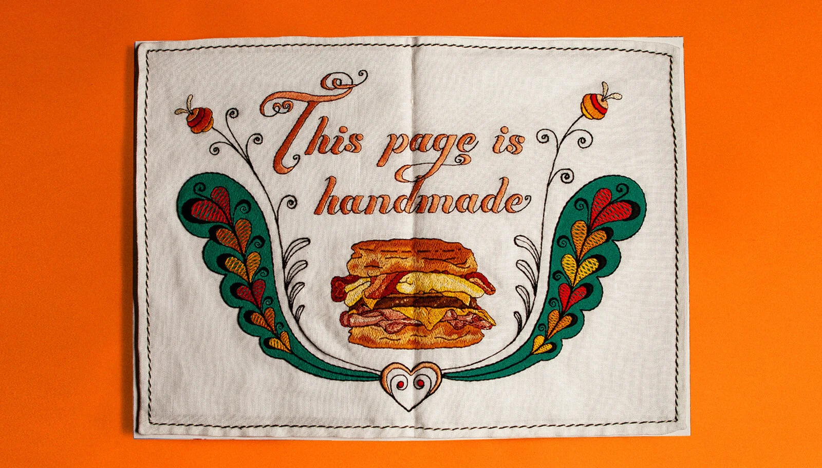 Embroidery artwork with the text "This page is handmade", a sandwich illustration, surrounded by colorful floral and leaf patterns on a white cloth background, set against an orange surface.