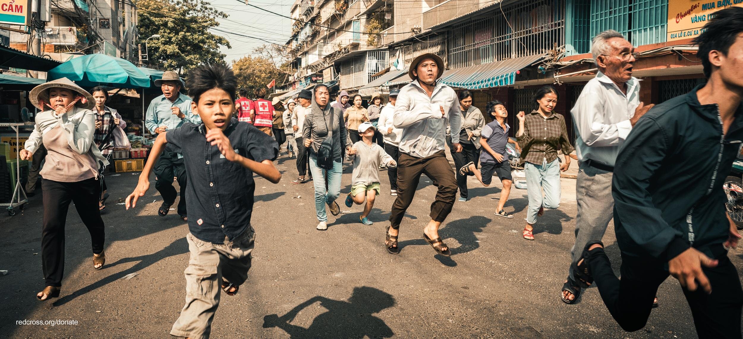 A group of people running on a street in an urban area, with some shopfronts and signs, and overhead wires in the background, during daytime.