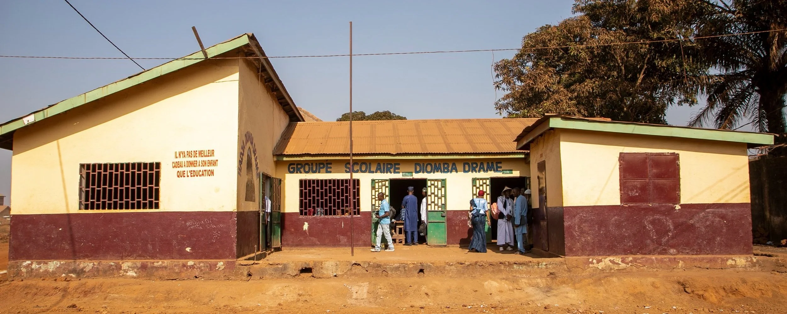 A small school building with a yellow and maroon exterior, crowded with students and teachers entering and exiting, located in a rural area with trees in the background.